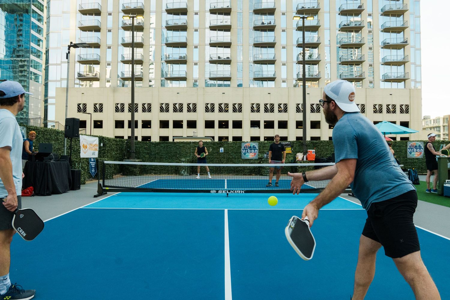 Two men playing pickleball on a blue outdoor court with a city high-rise building in the background.
