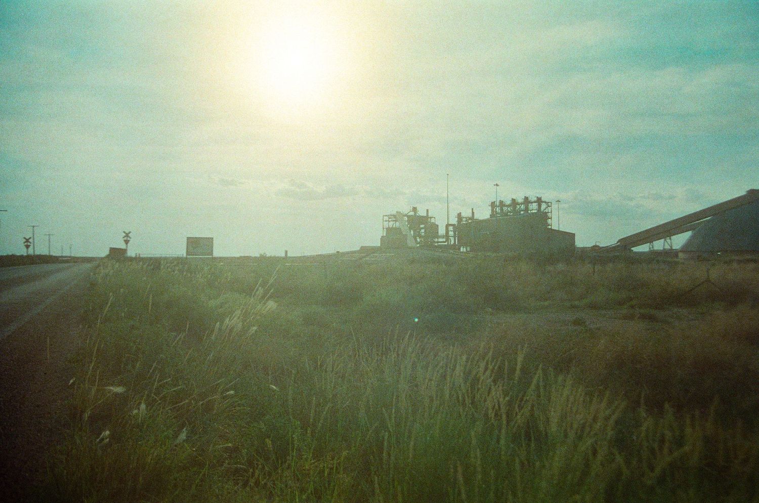 Industrial facility silhouetted against bright sun over green field on overcast day.