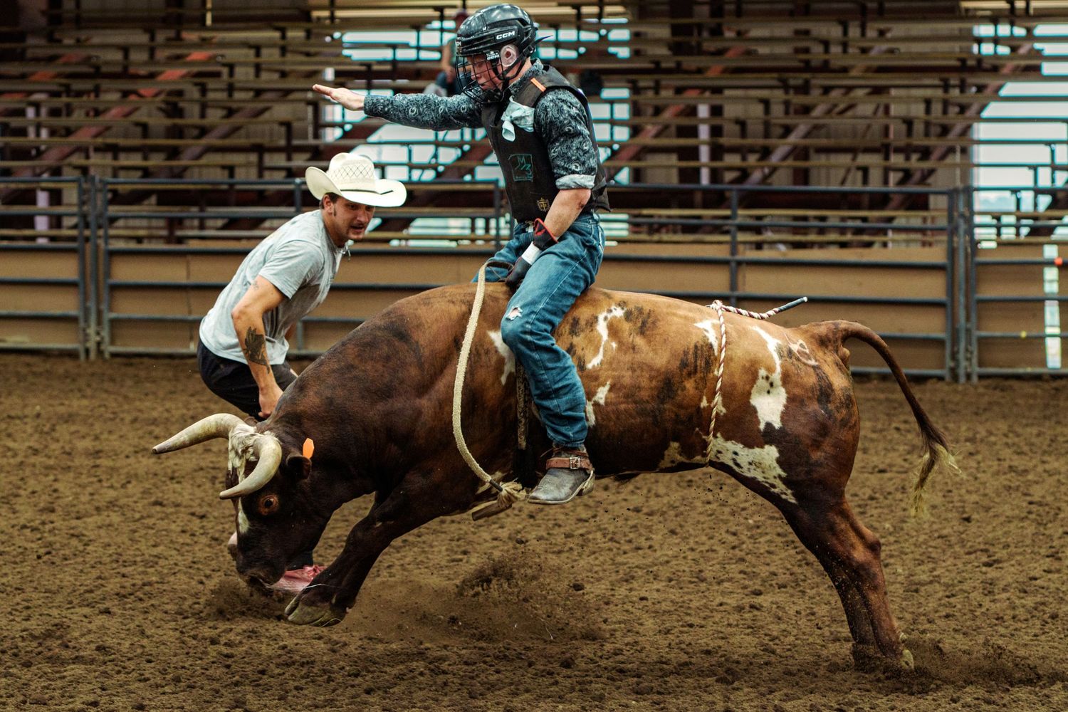 A bull rider grips tightly as a brown bull bucks at an indoor rodeo arena while a rodeo clown assists nearby.