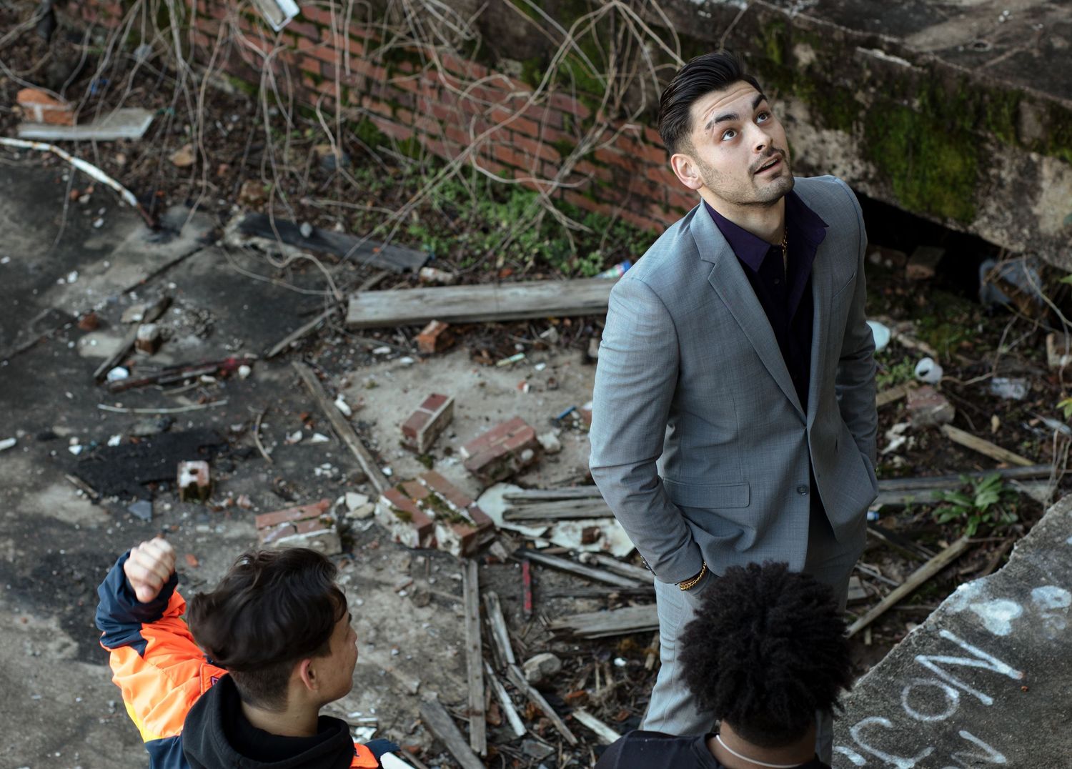 Man in gray suit looks up at damaged brick building exterior with children nearby in urban setting.