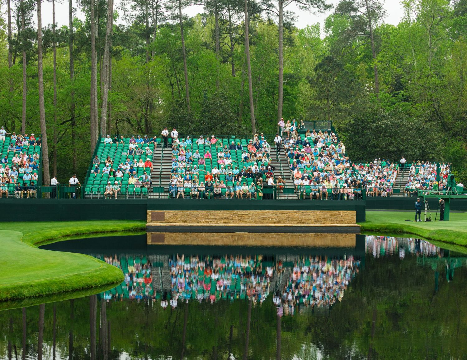 Spectators fill green grandstands beside a reflective pond at Augusta National Golf Club during The Masters tournament.
