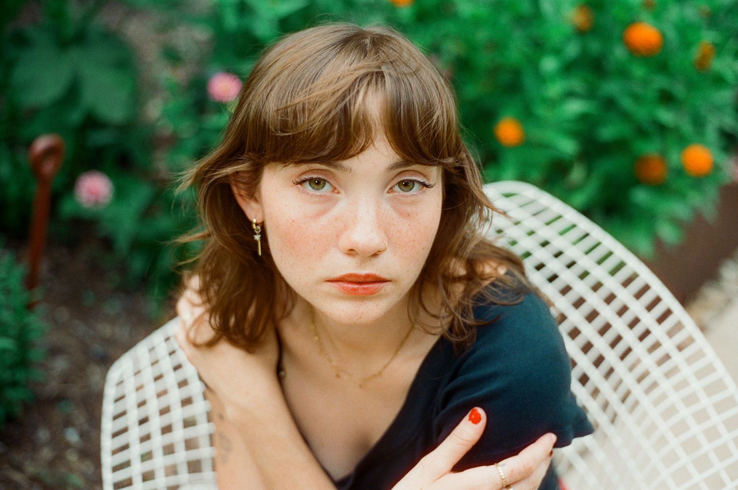 Young woman with freckles and bangs sits in garden chair outdoors.