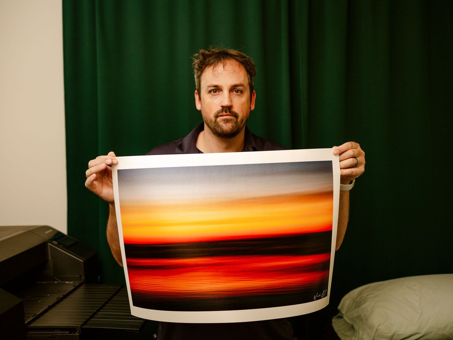Man displaying a vibrant abstract sunset fine art print against a green curtain backdrop.