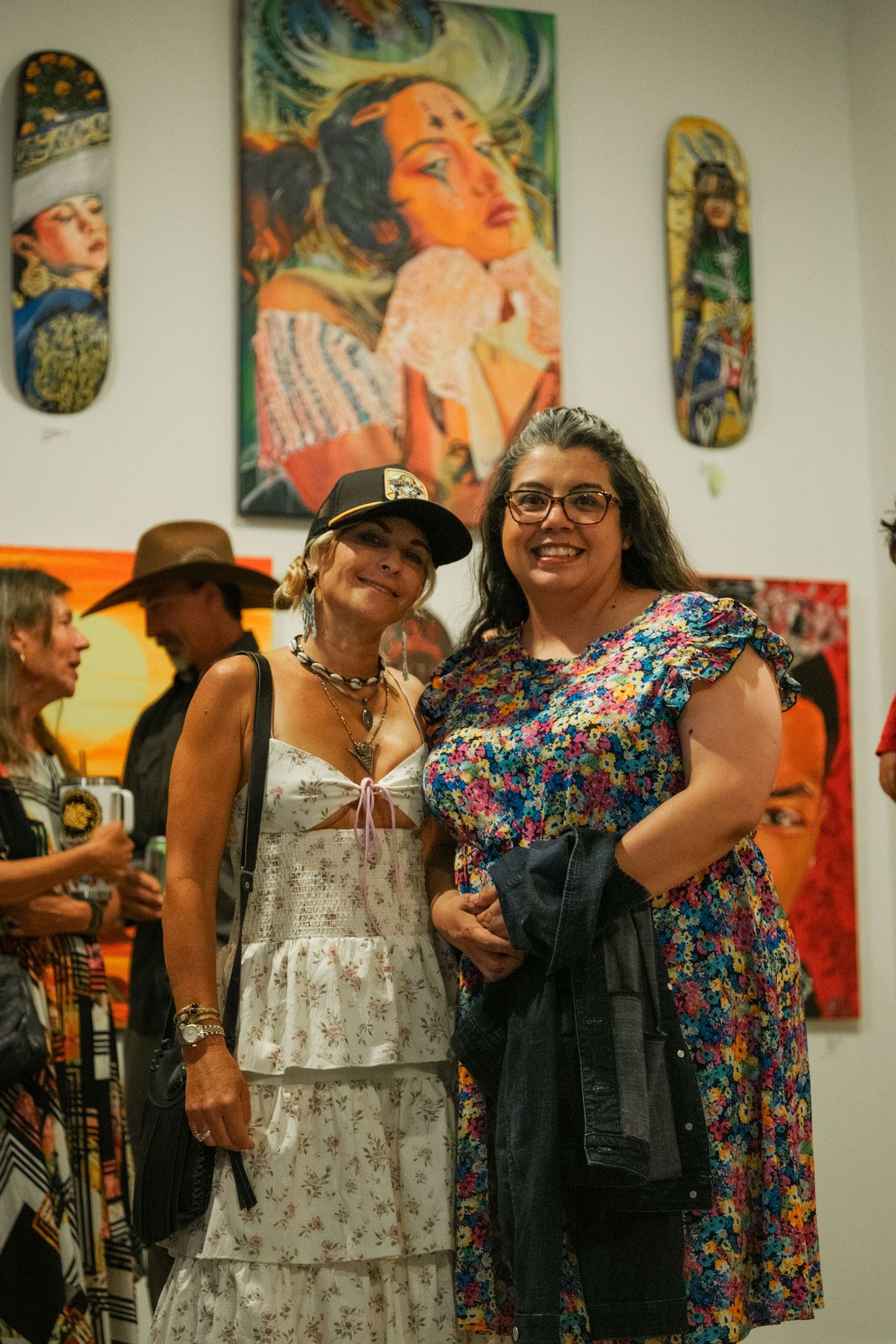 Two women smiling together in front of colorful figurative paintings at an art gallery exhibition.