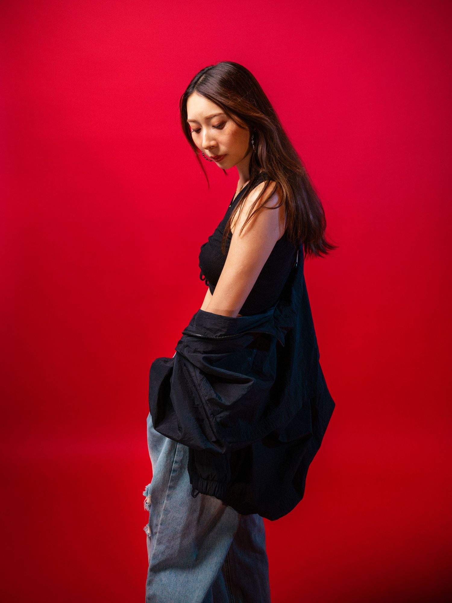 Asian woman in black crop top and ripped jeans looks down gracefully against a vibrant red backdrop.
