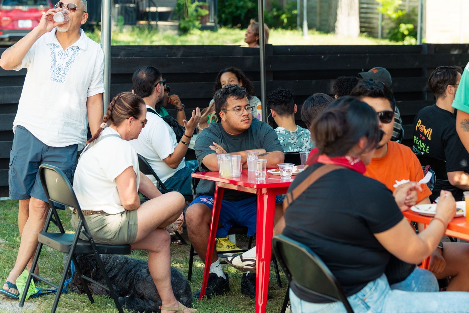 Diverse group of people dining outdoors at red tables on folding chairs at a casual event.