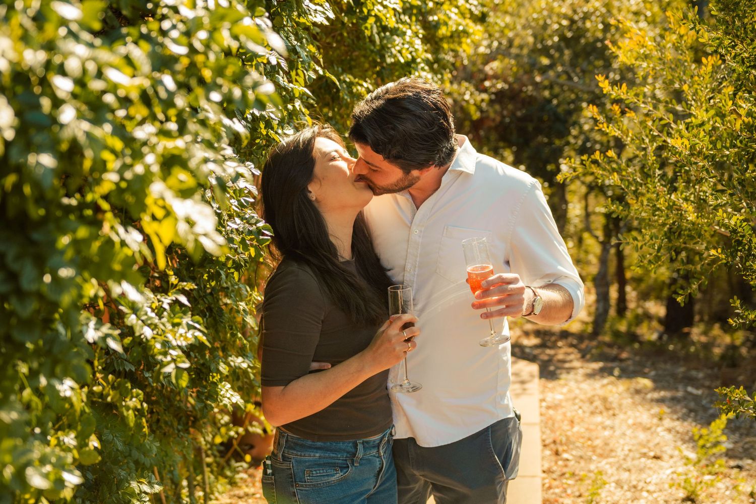 Couple kissing in a sunny vineyard, holding wine glasses, surrounded by lush green vines.