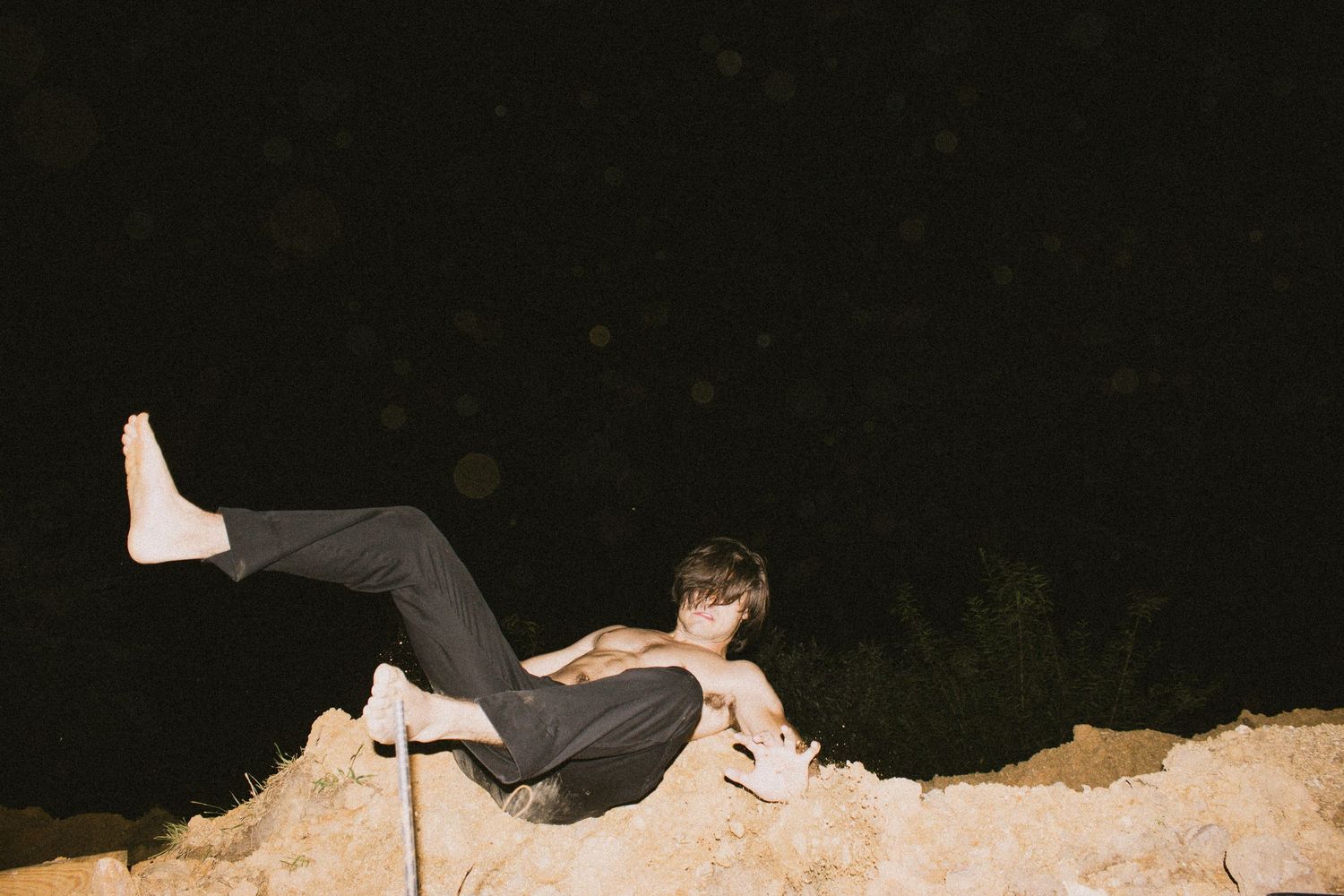 Person in black pants poses dynamically on sandy terrain at night, barefoot with one leg raised.