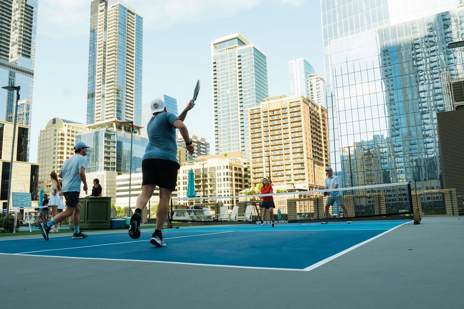 Players enjoy a rooftop tennis match on a blue court surrounded by modern city skyscrapers on a sunny day.