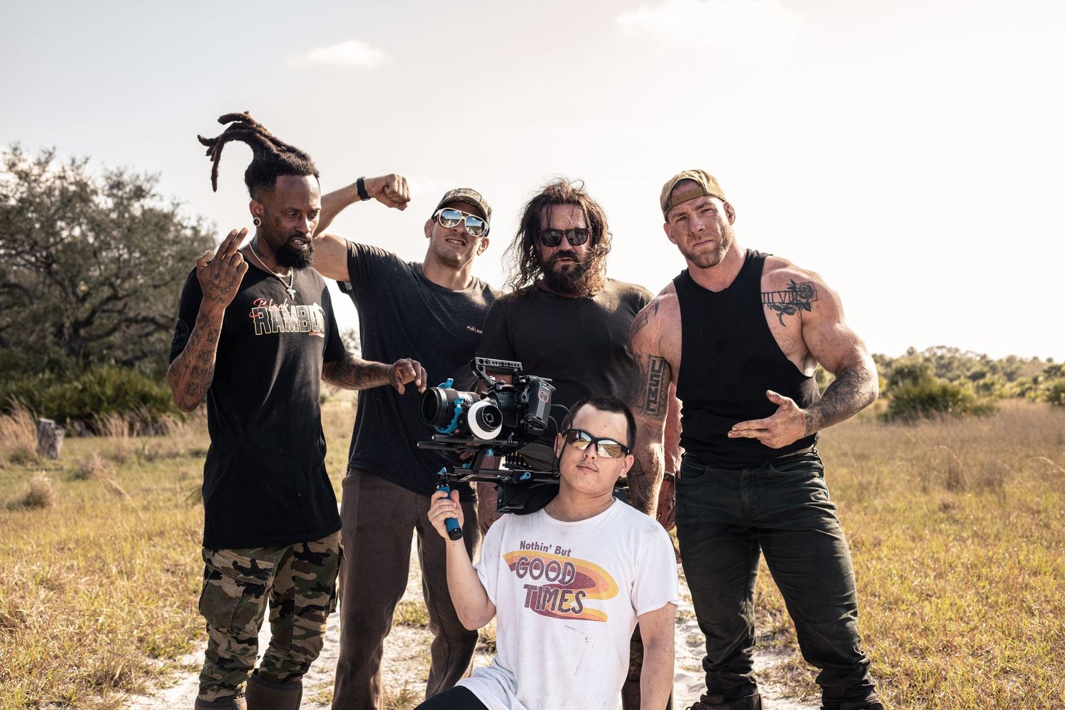 Group of five people posing outdoors with a camera rig in a sunny field, smiling and flexing.