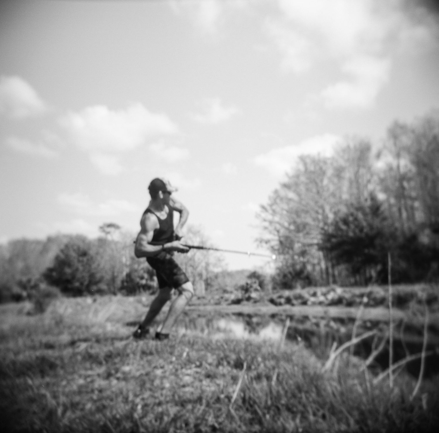 Black and white photo of a boy fishing by a pond in a rural landscape with trees and cloudy sky.