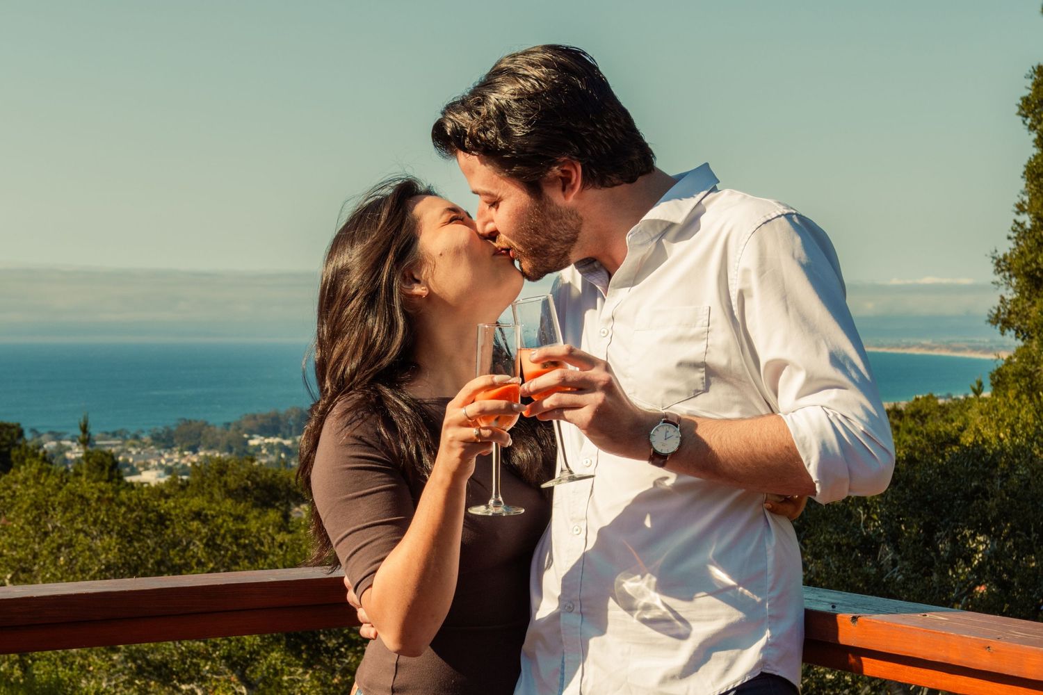 Romantic couple kissing and clinking wine glasses on a scenic overlook with ocean views on a sunny day.