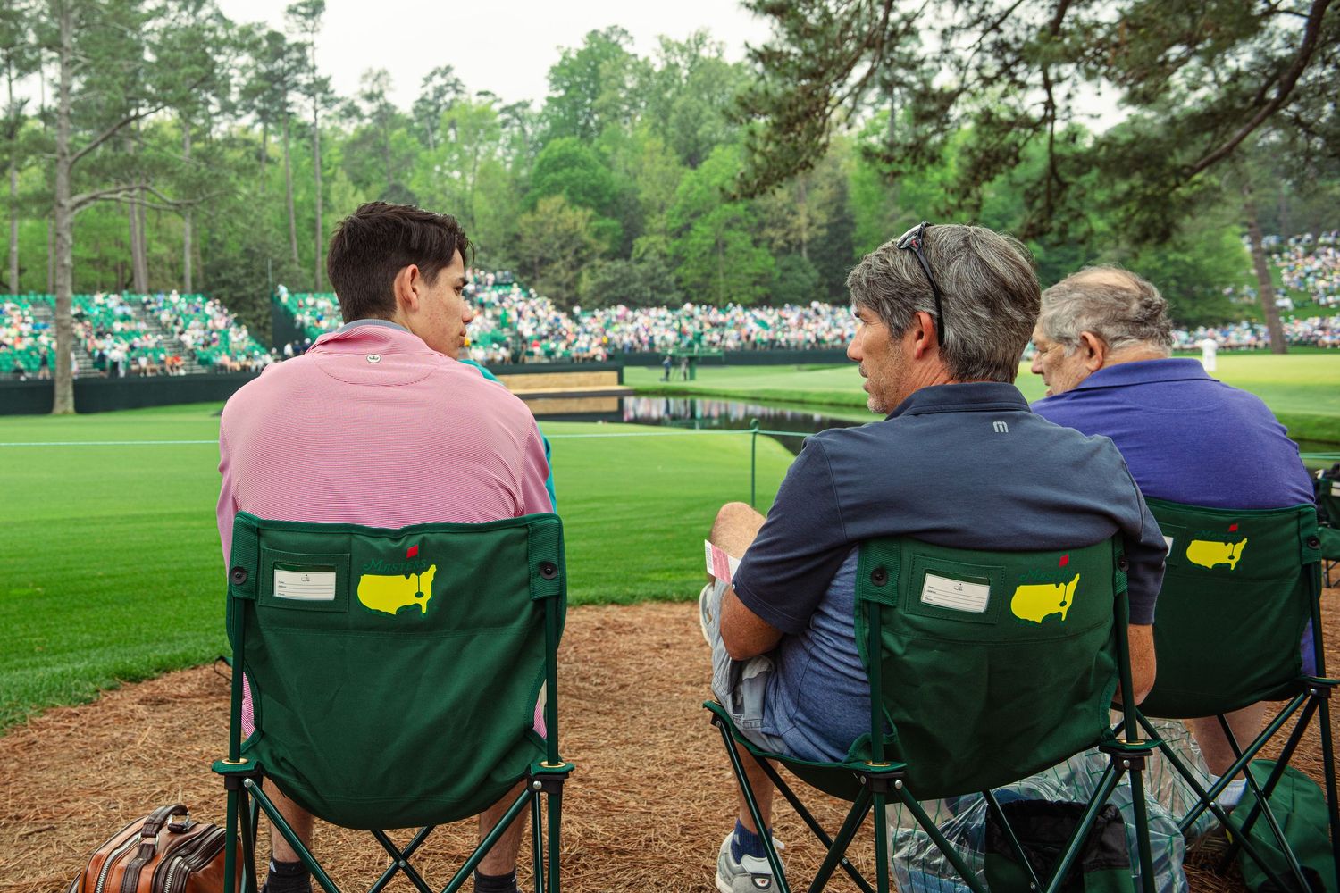 Three spectators sit in green Masters Tournament chairs watching golf at Augusta National Golf Club.