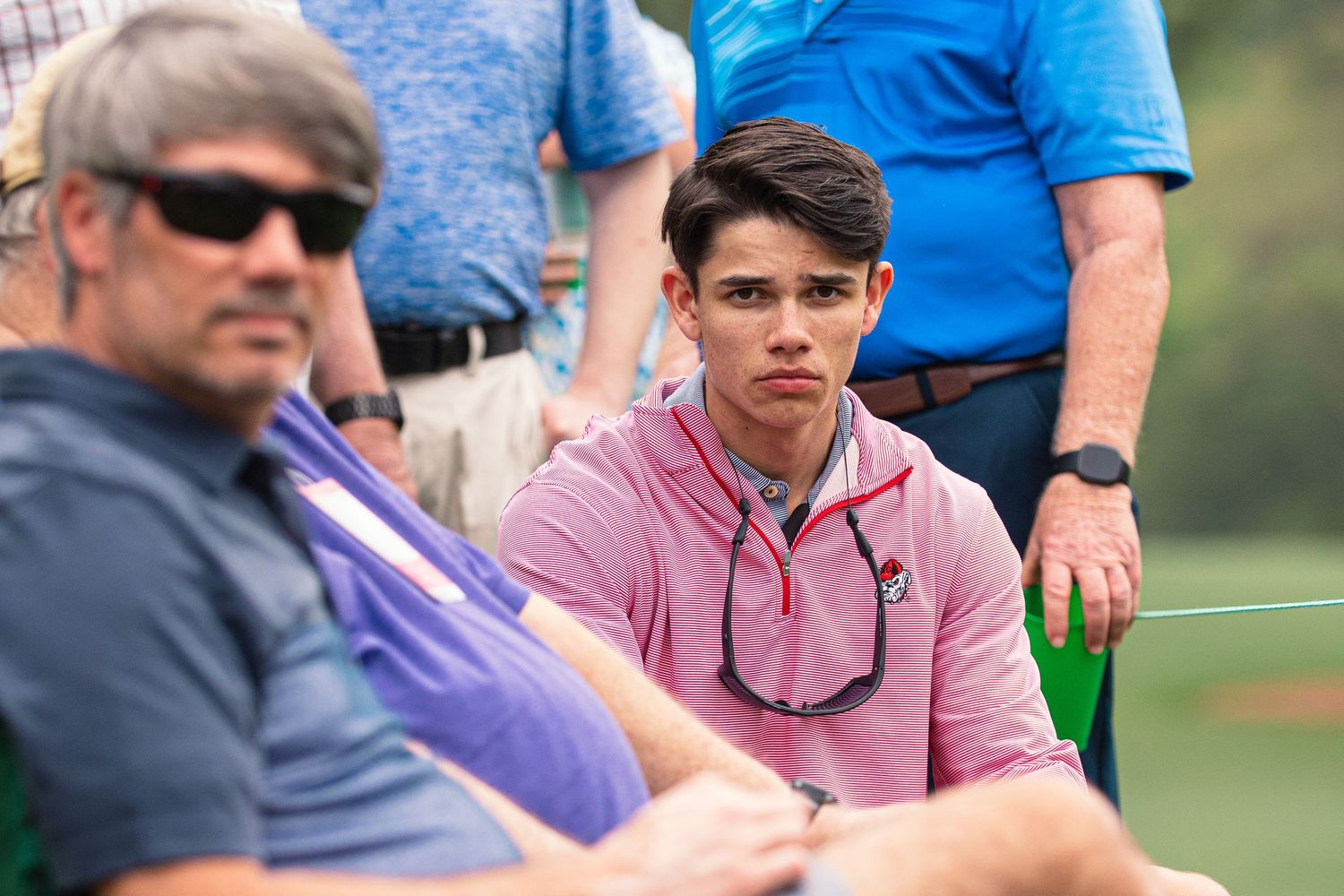 Young man in pink polo shirt sitting with older man in sunglasses at an outdoor golf event.
