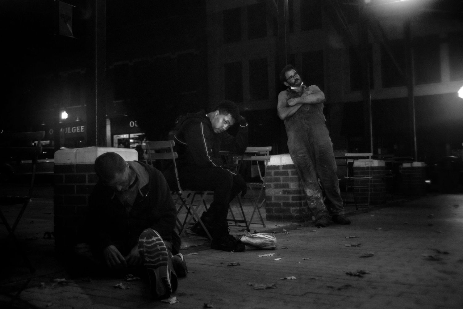Three men gathered at night in a dimly lit urban setting, captured in black and white photography.