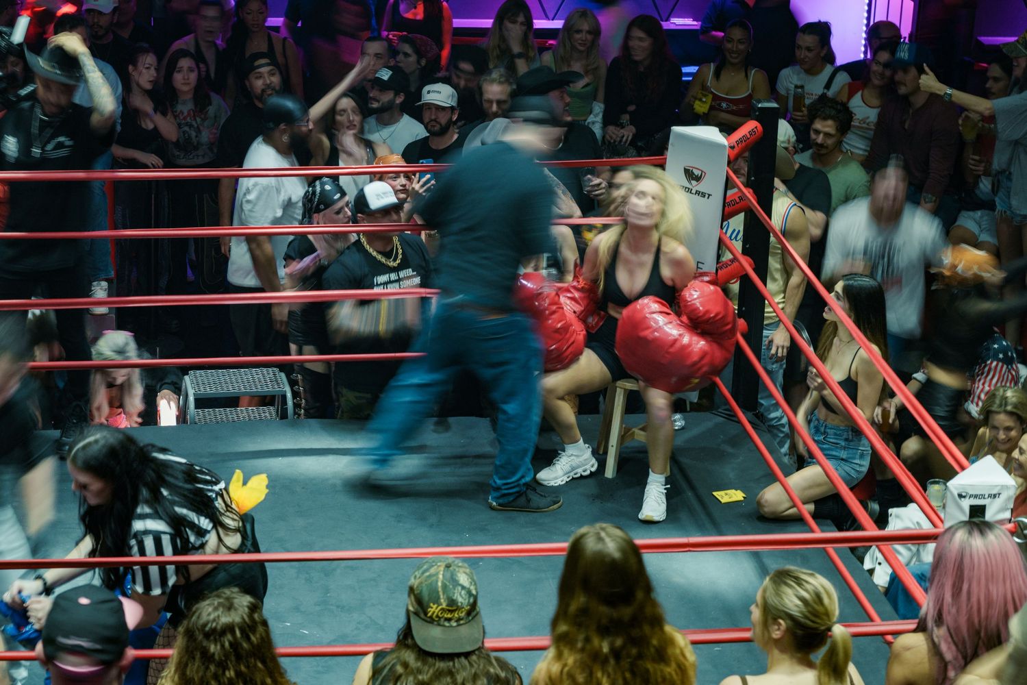 A female boxer in red gloves sits in the corner of a boxing ring as a referee approaches during a match.