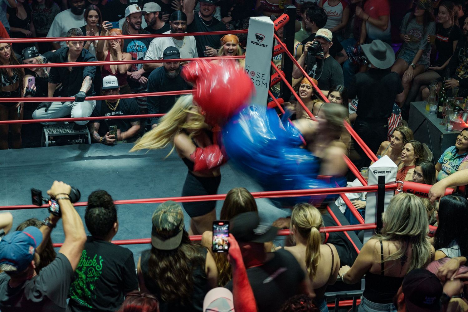 Two female boxers in oversized red and blue gloves exchange powerful punches in a crowded boxing ring.