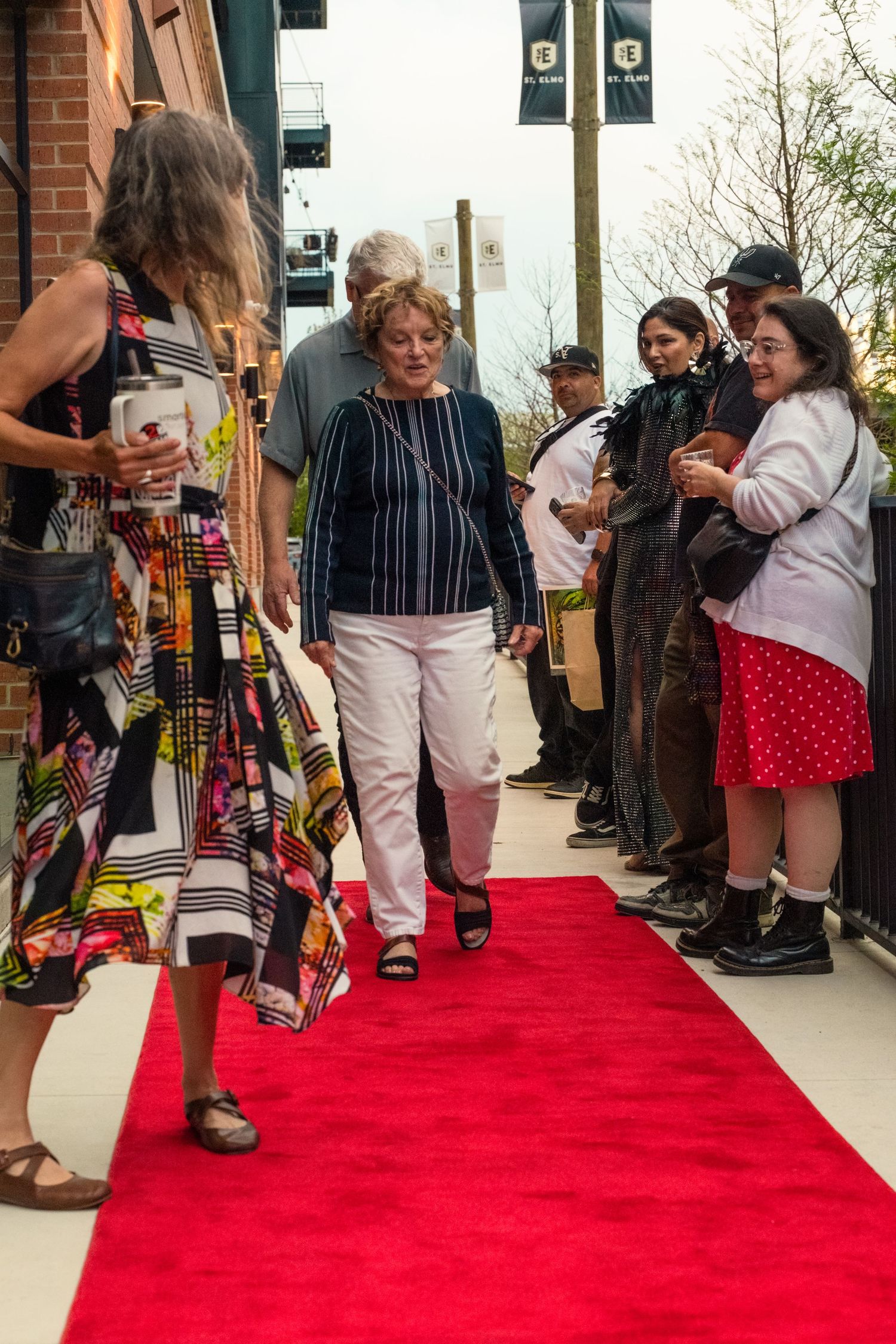 Guests walking on a red carpet at an outdoor event entrance with crowd of onlookers lining the sides.
