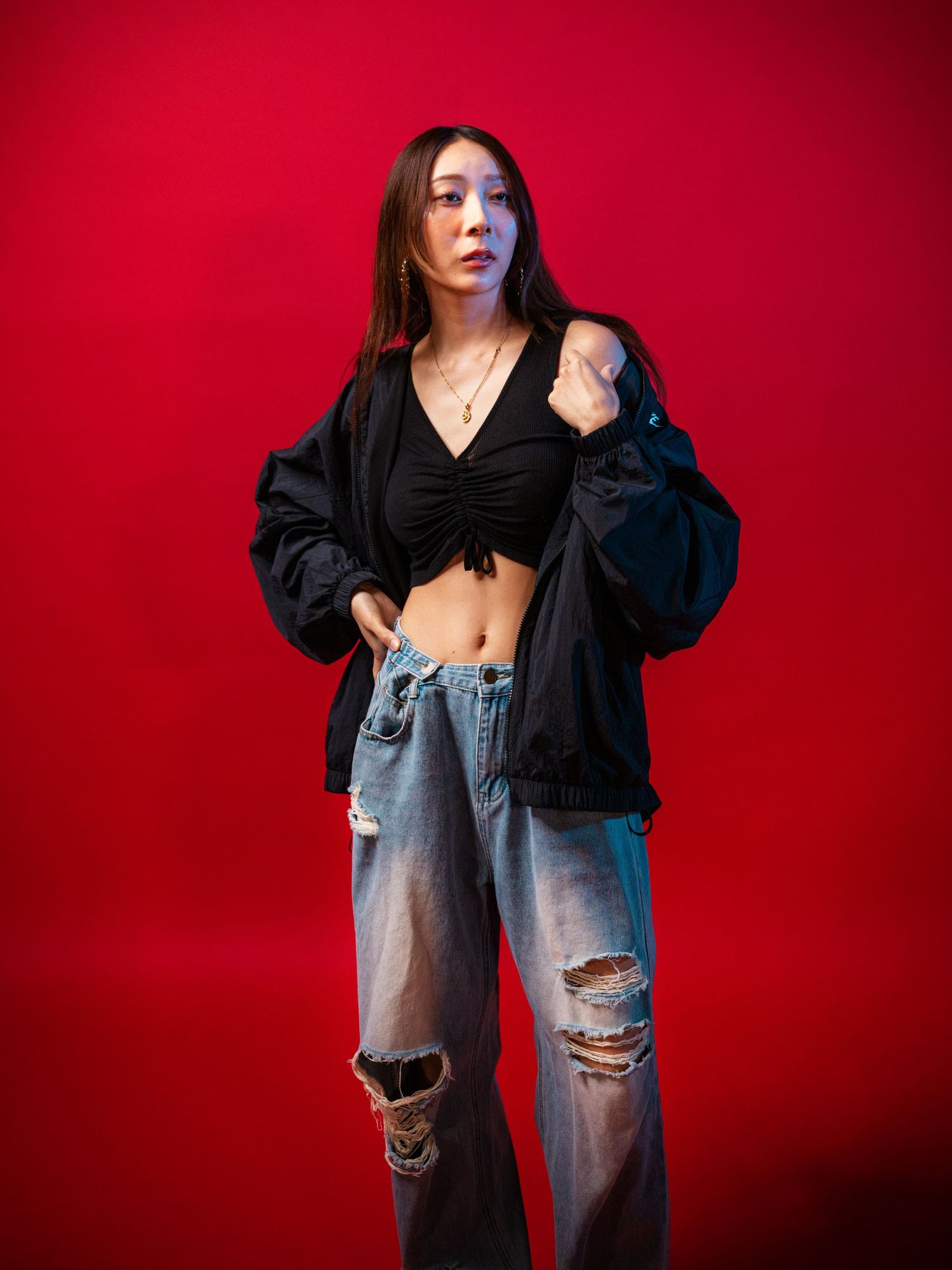 Asian woman in black crop top and ripped jeans poses confidently against a bold red studio background.
