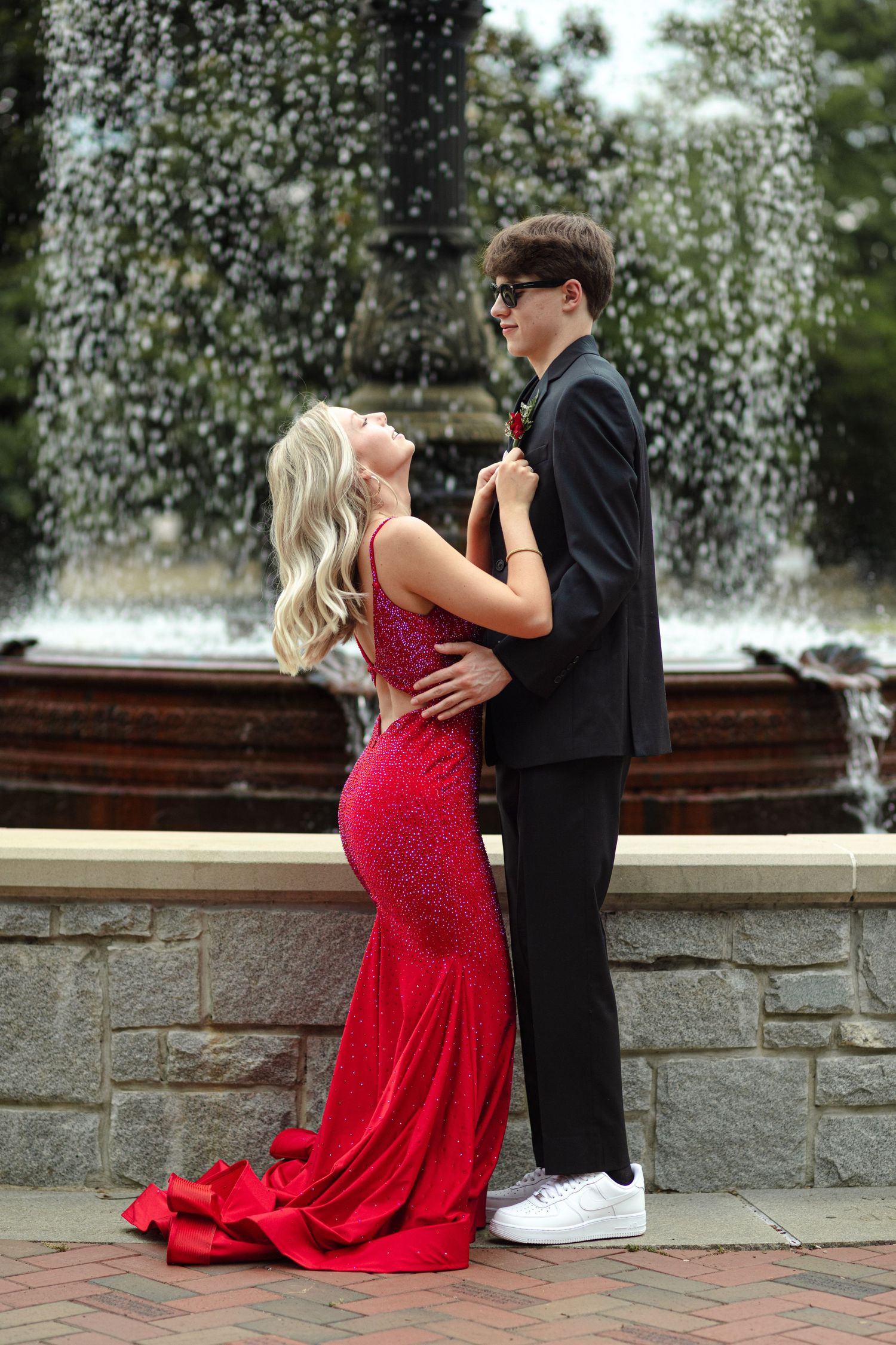 Couple posing in formal prom attire in front of a fountain, woman in red sequin gown and man in black suit.