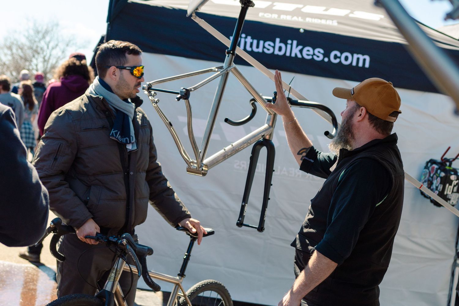 Two men discussing a titanium bicycle frame at an Itus Bikes outdoor expo booth on a sunny day.