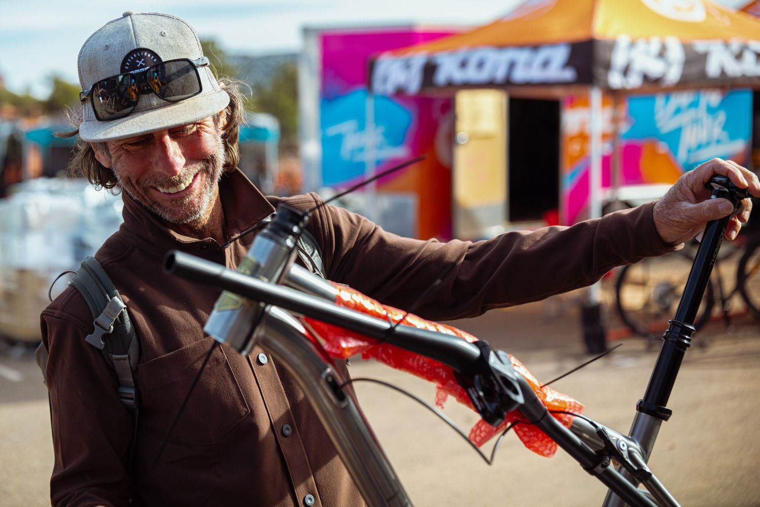 Smiling man in brown jacket and cap holding a red Kona mountain bike at an outdoor cycling event.