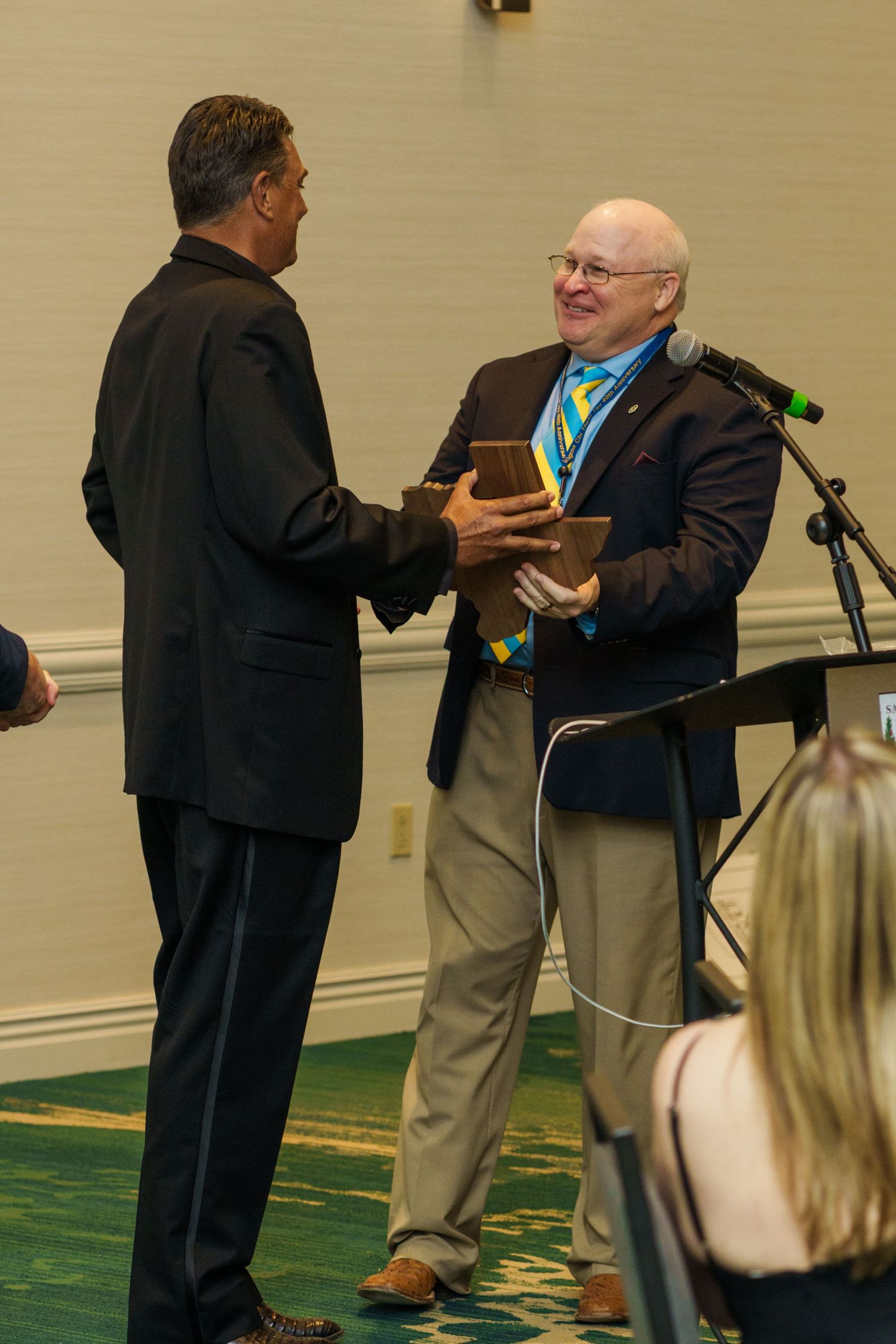 Two men shaking hands at an awards ceremony, one presenting a wooden plaque in a hotel ballroom.