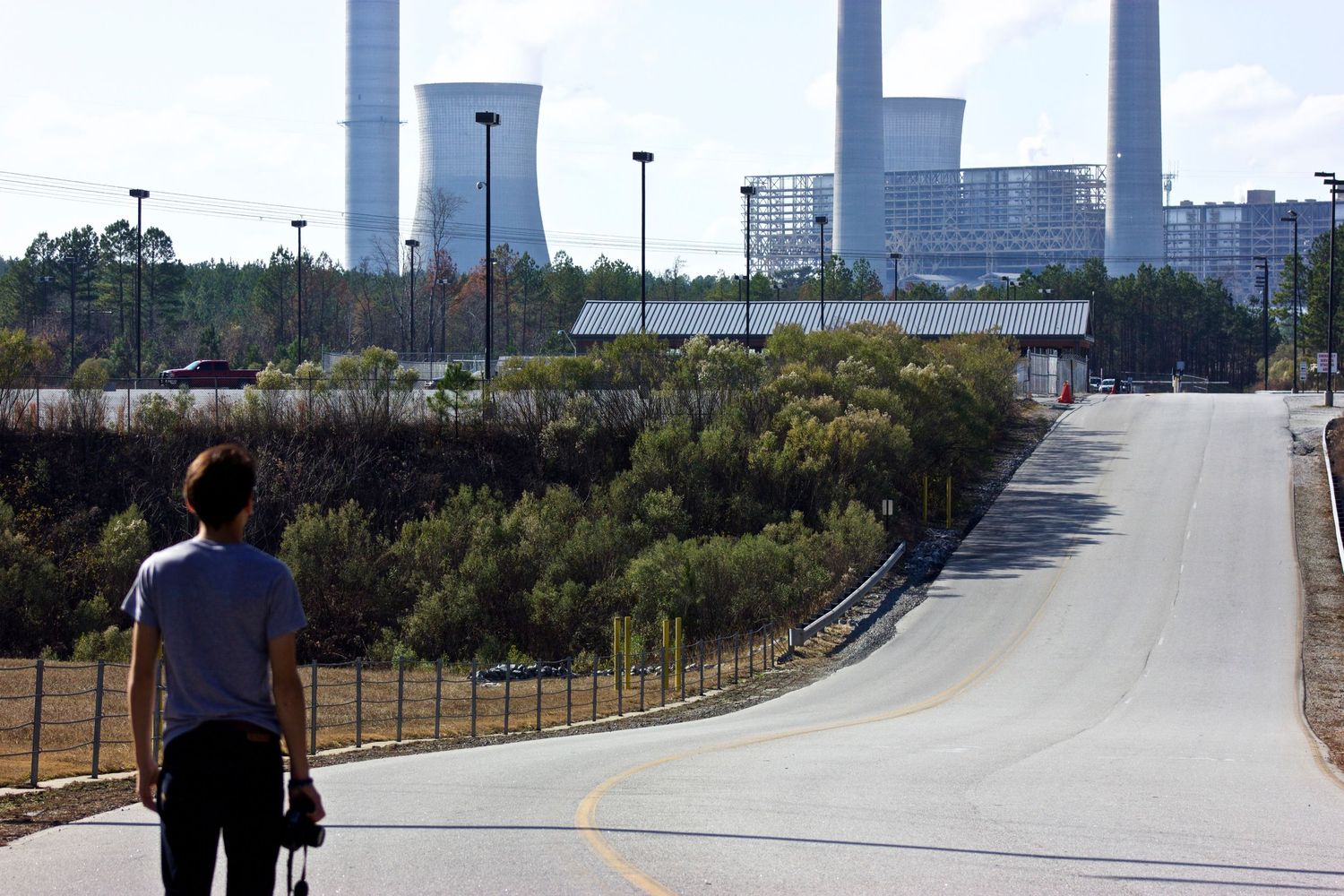 Person standing on empty road leading to industrial power plant with cooling towers against clear sky.