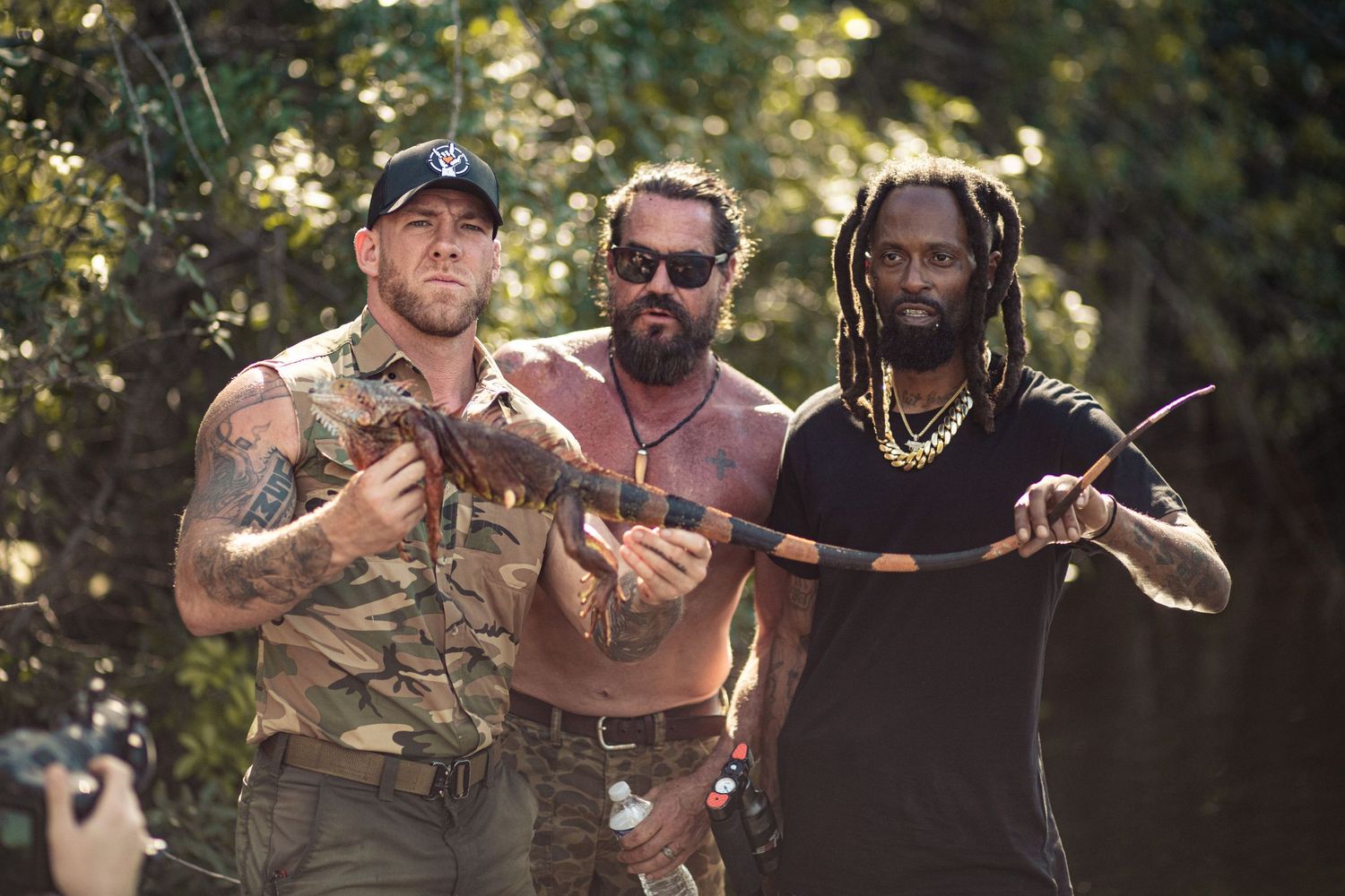 Three men posing outdoors in jungle setting, holding weapons and traditional bow, wearing camouflage and casual attire.