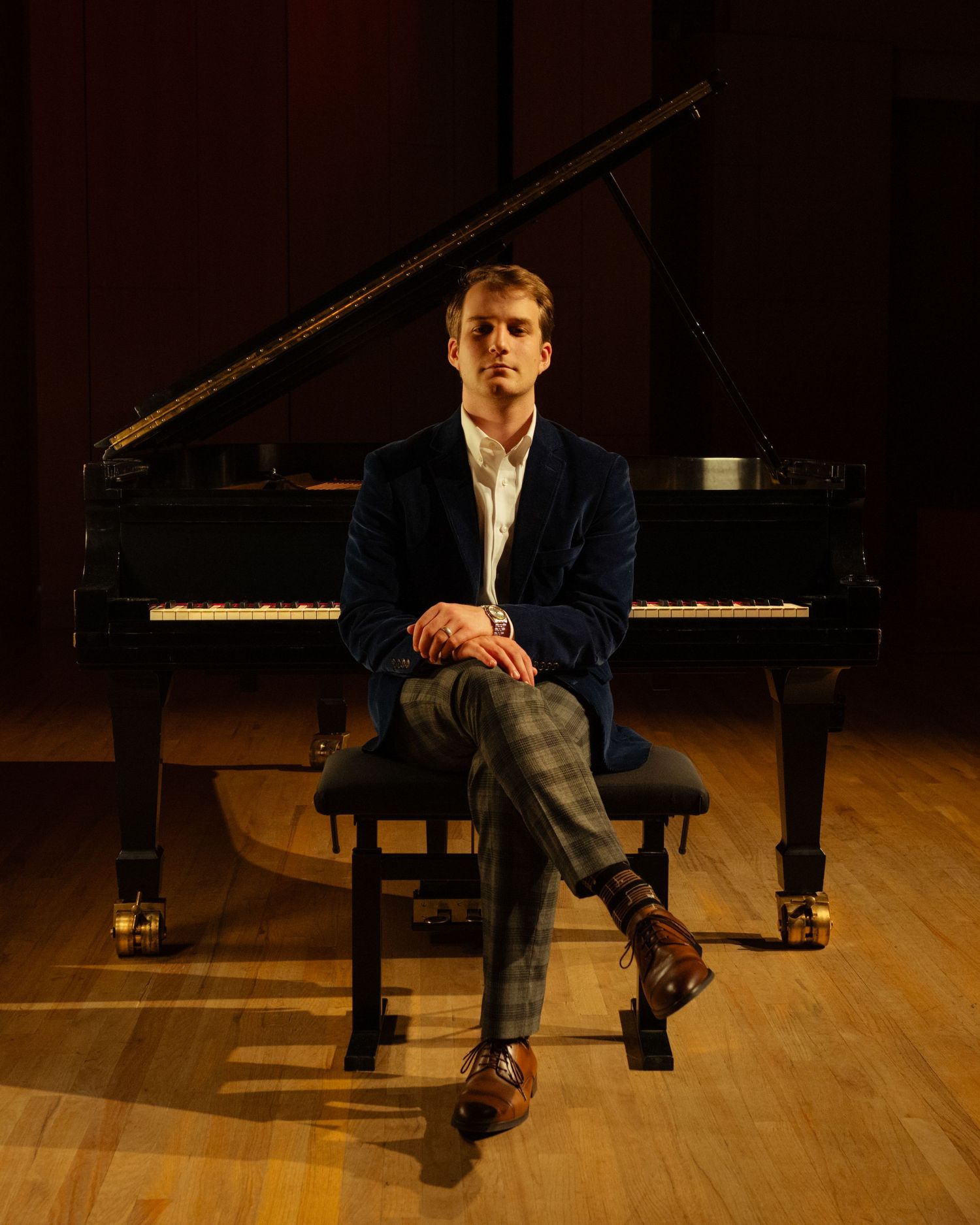 Young man in navy blazer sits before grand piano on concert stage.