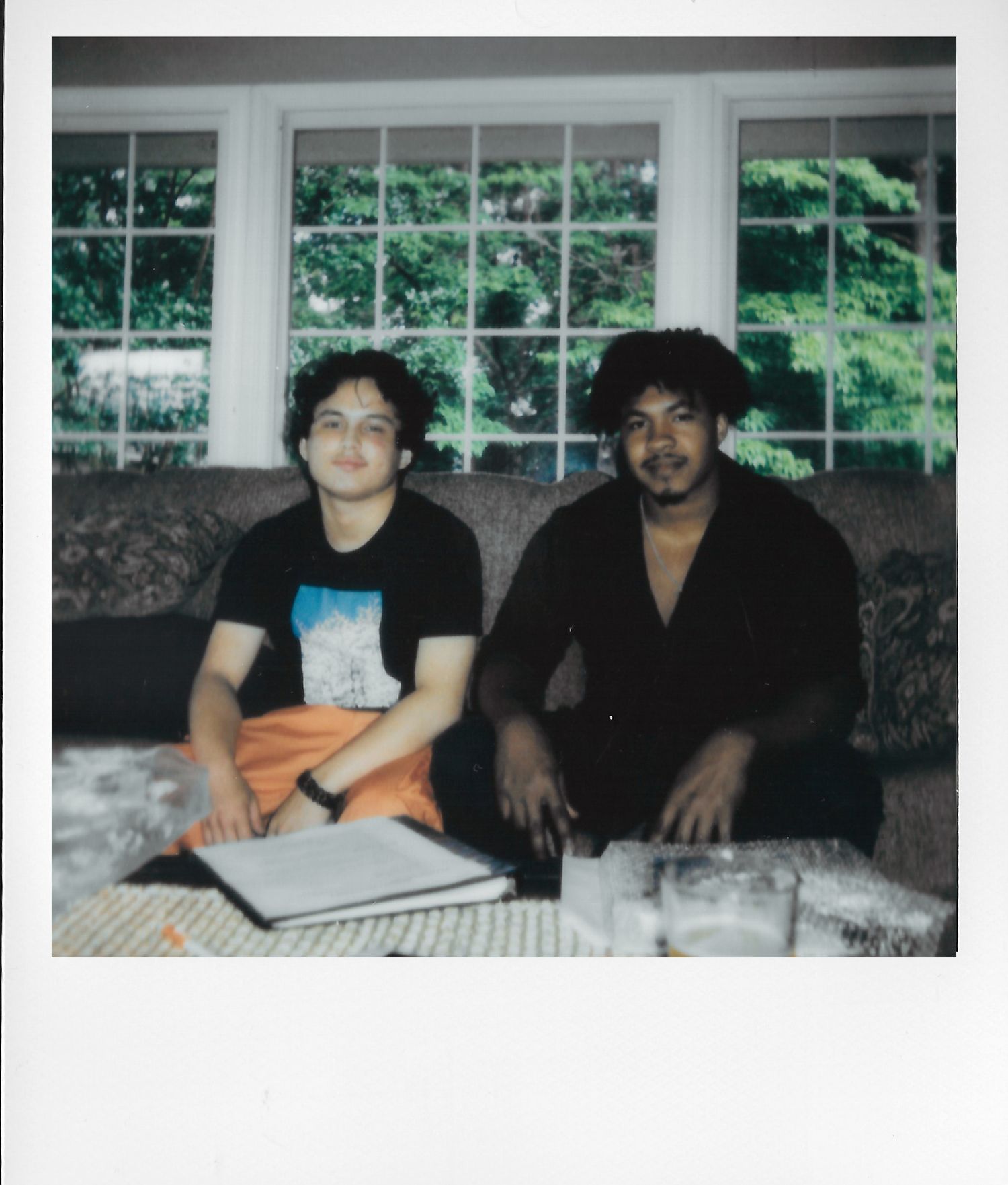 Two people sitting on a couch indoors, posing for a Polaroid-style photo with large windows and greenery behind them.