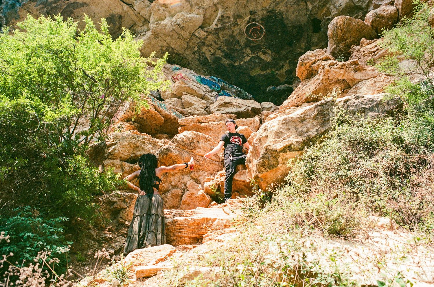 Two people exploring rocky terrain surrounded by lush green trees in a scenic outdoor canyon setting.