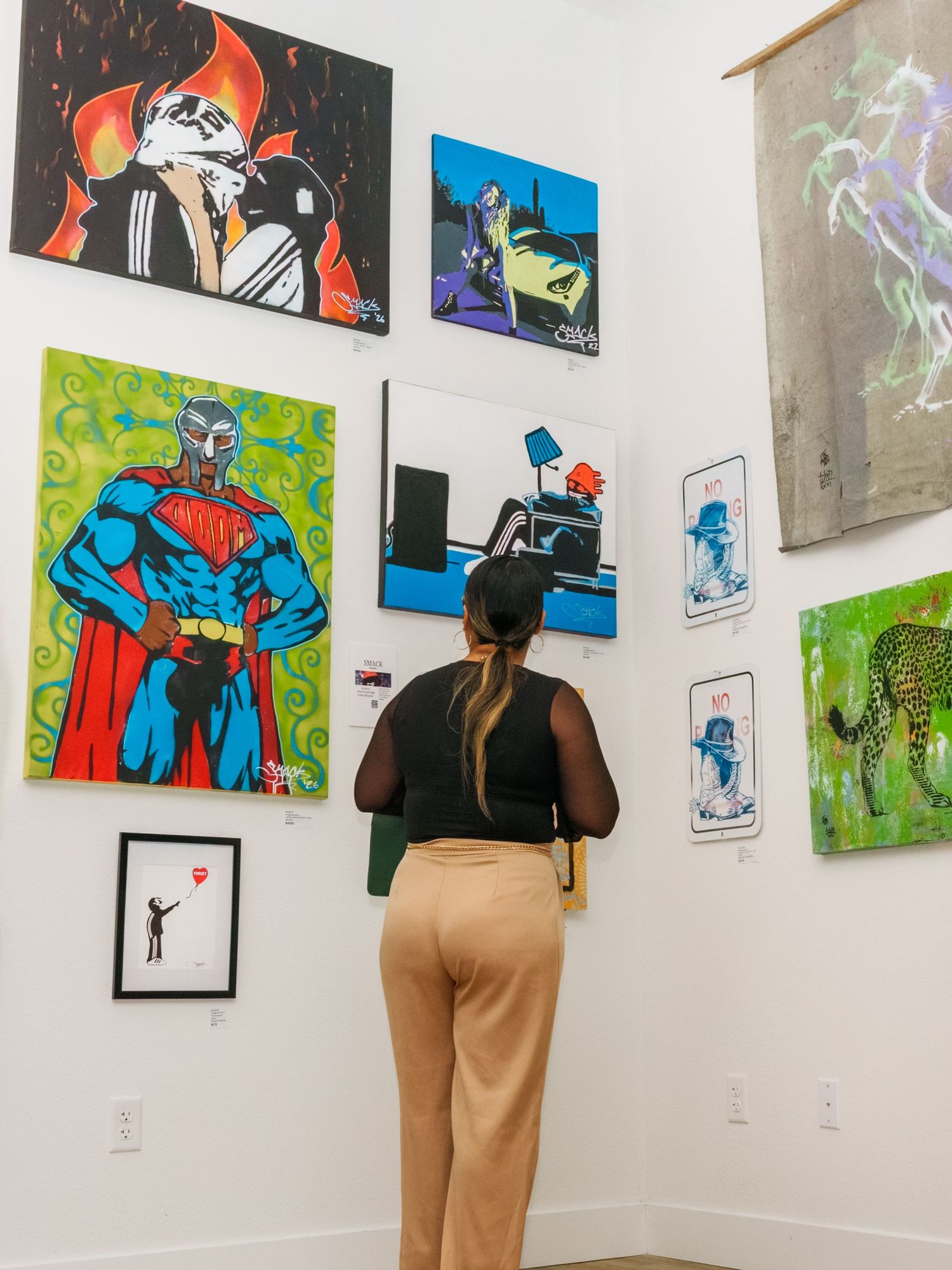 Woman viewing colorful pop art and street art paintings displayed on white gallery wall.