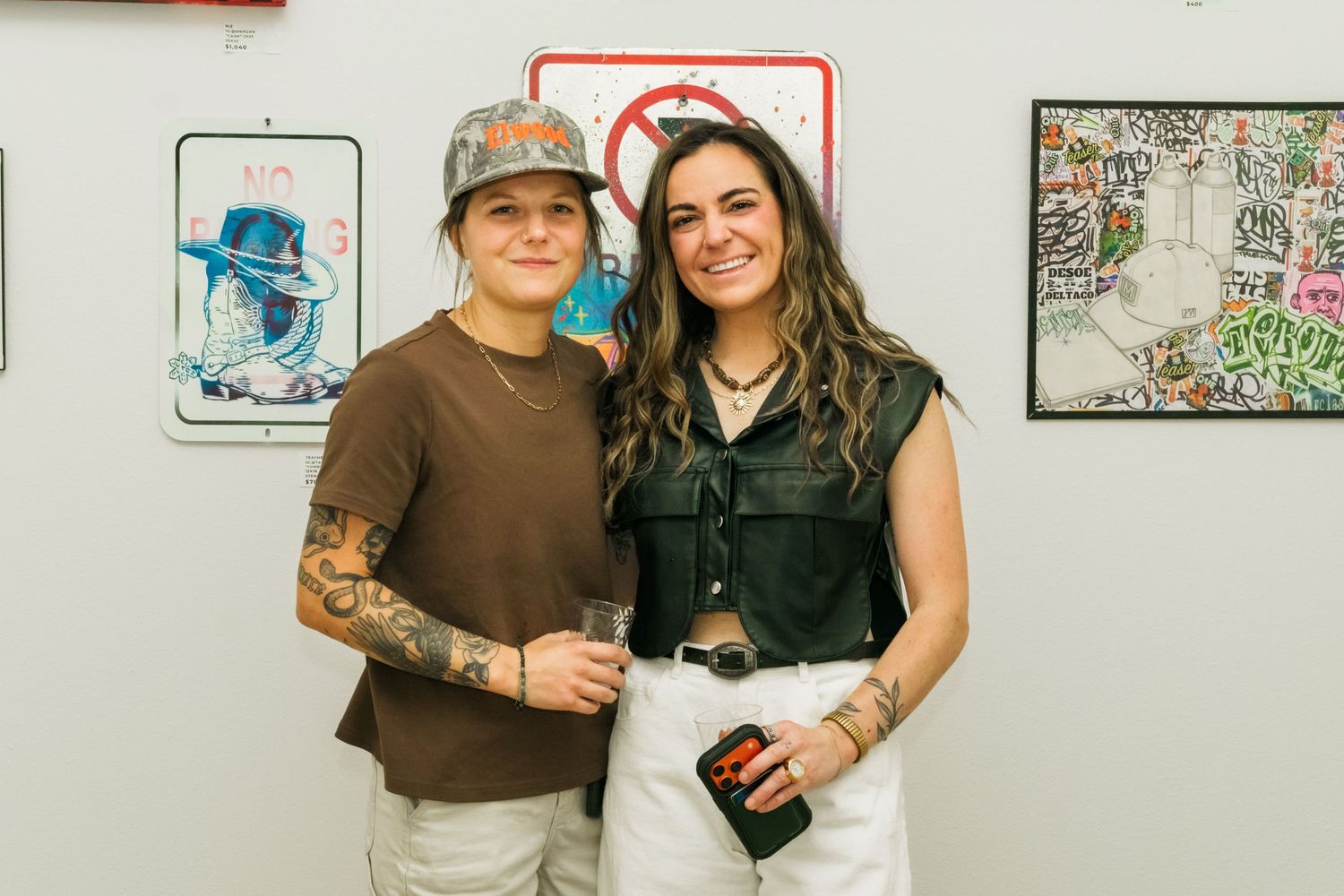 Two tattooed women smile together at an art gallery opening surrounded by street art and graffiti-style paintings.