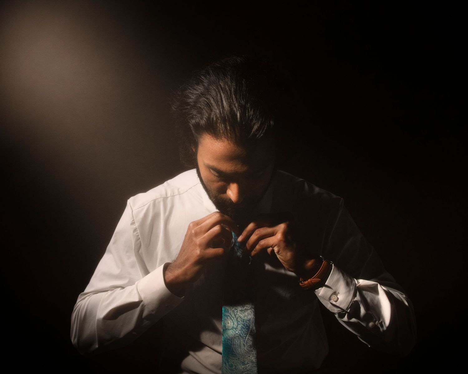 Dramatic portrait of a man in white shirt adjusting tie under a single spotlight in darkness.