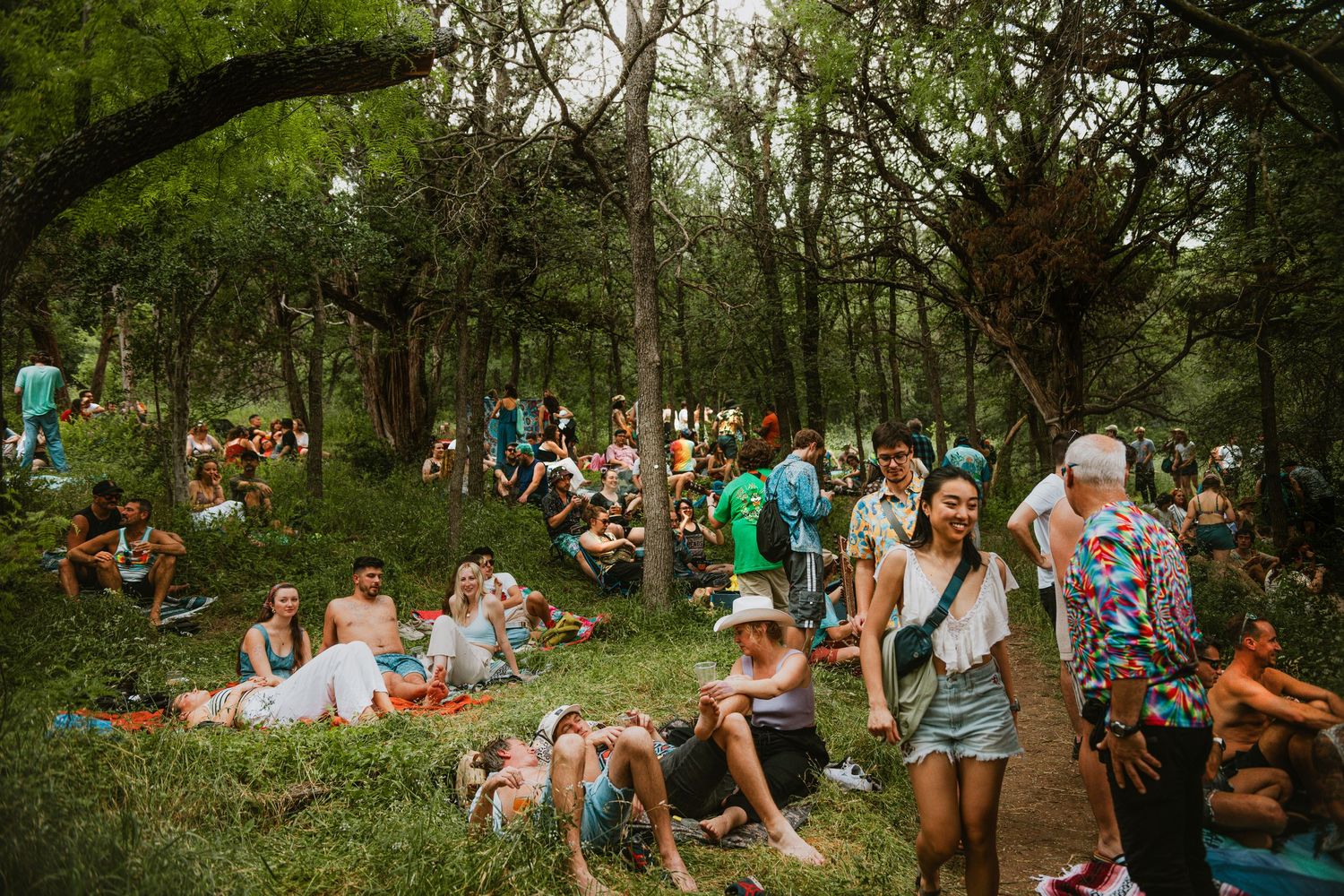 Crowds of festival-goers relaxing on grass under lush green trees at an outdoor music festival called Eyore's Birthday