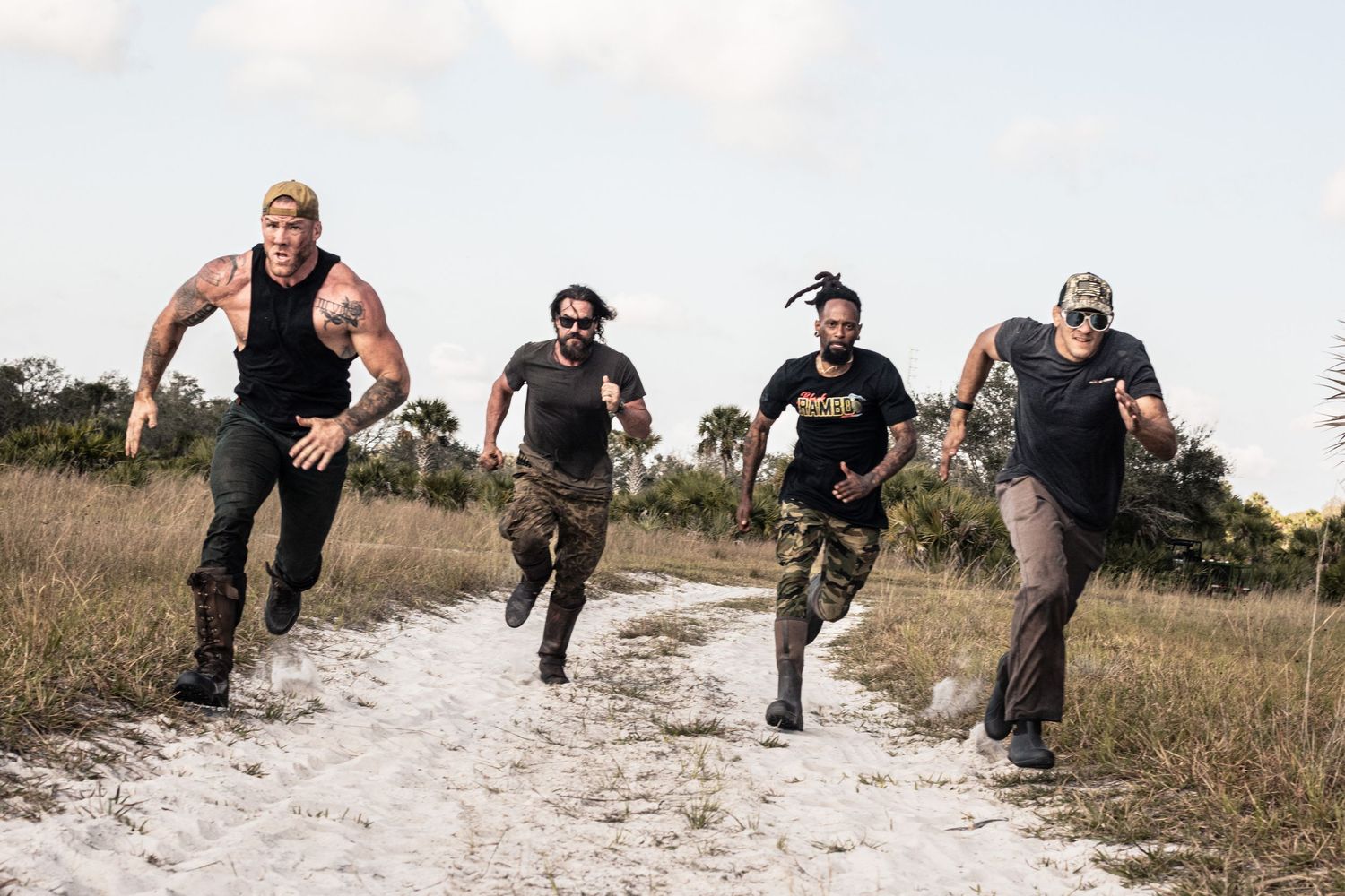 Four fit men sprinting down a sandy path surrounded by tall grass in athletic and camouflage clothing.