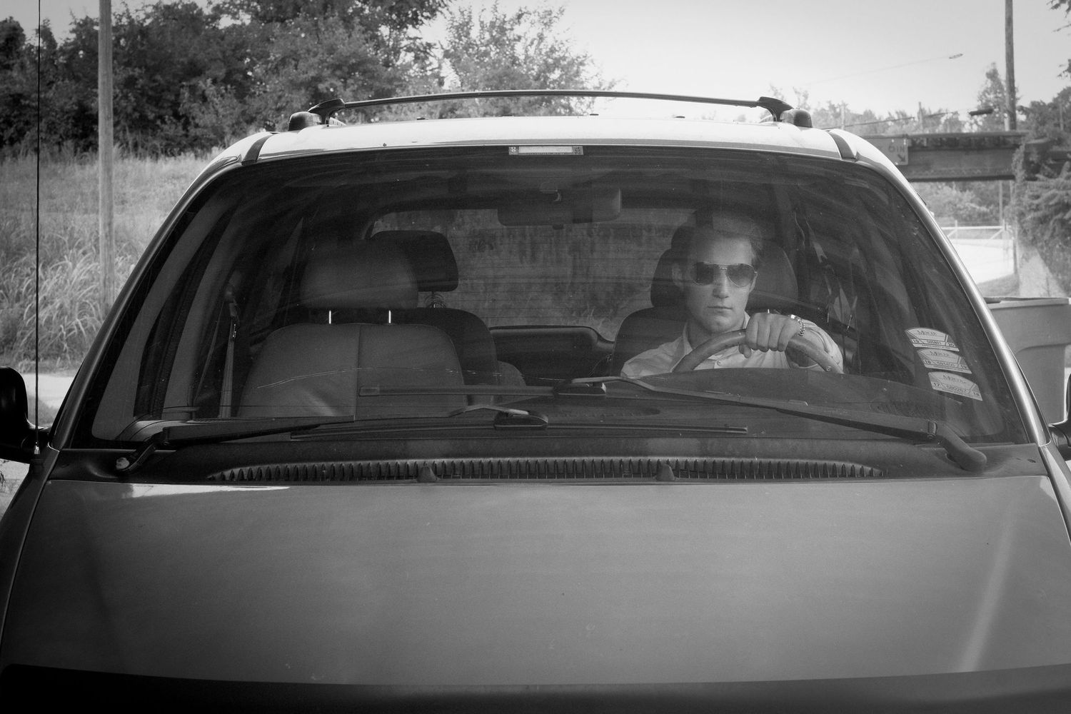 Black and white photo of a person wearing sunglasses driving an SUV, viewed through the windshield.