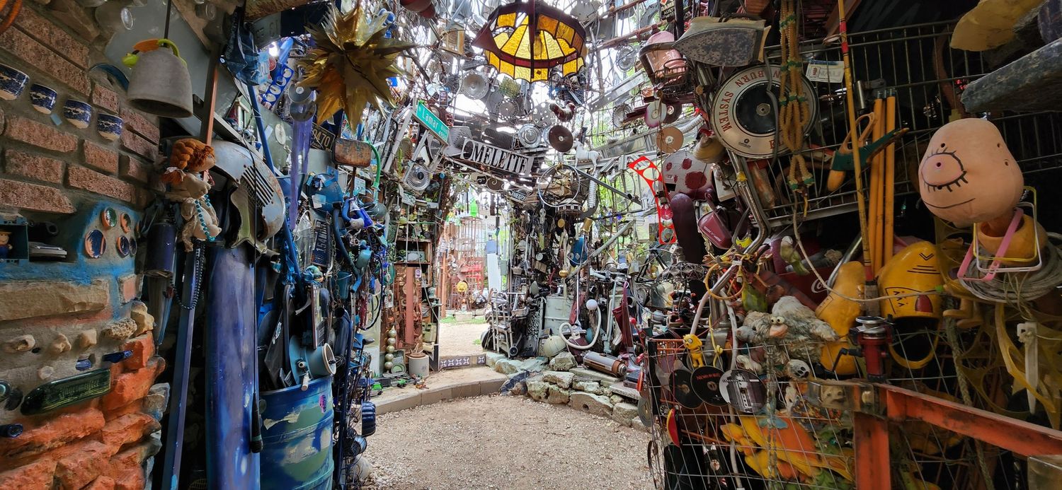 A narrow alleyway filled with colorful eclectic art installations, sculptures, and found objects creating an immersive outdoor gallery.