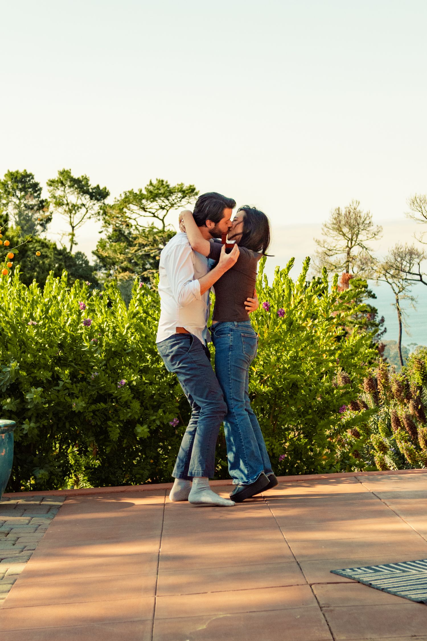 A couple sharing a romantic kiss outdoors on a patio surrounded by lush green trees and gardens.