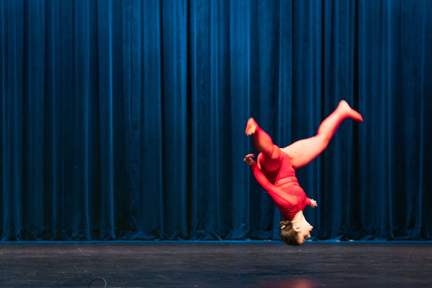 Dancer in red outfit performing a one-handed handstand on stage with blue curtain backdrop.