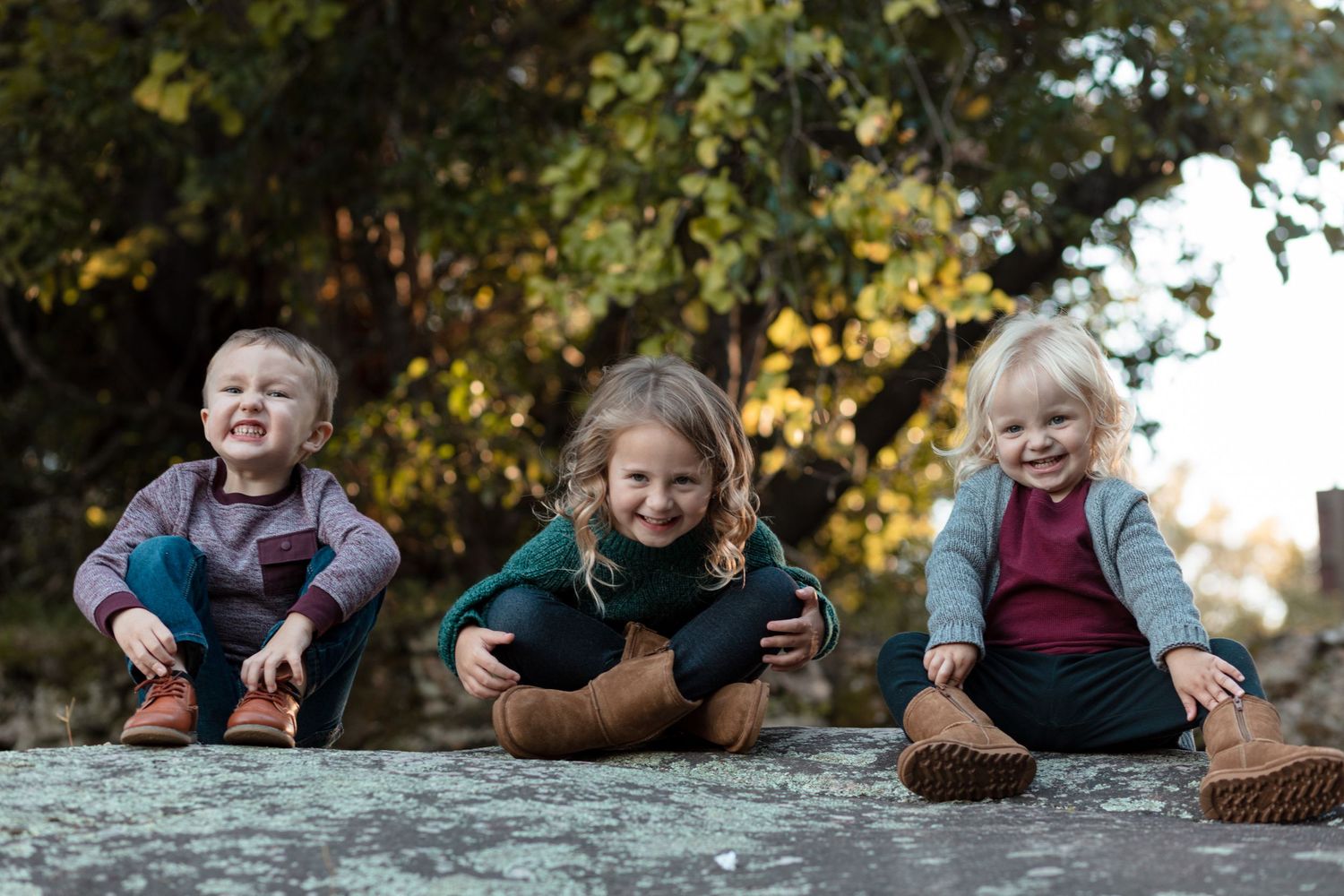 Three young children sitting on a rock outdoors, smiling in warm autumn sunlight.