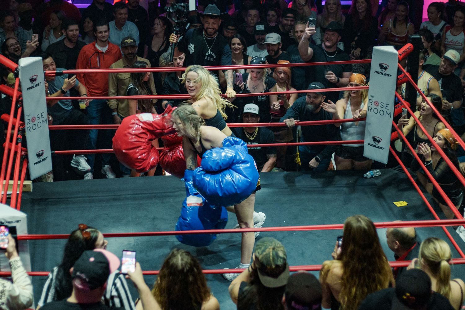 Two female boxers in red and blue oversized gloves grapple in a ring before an energetic live audience.