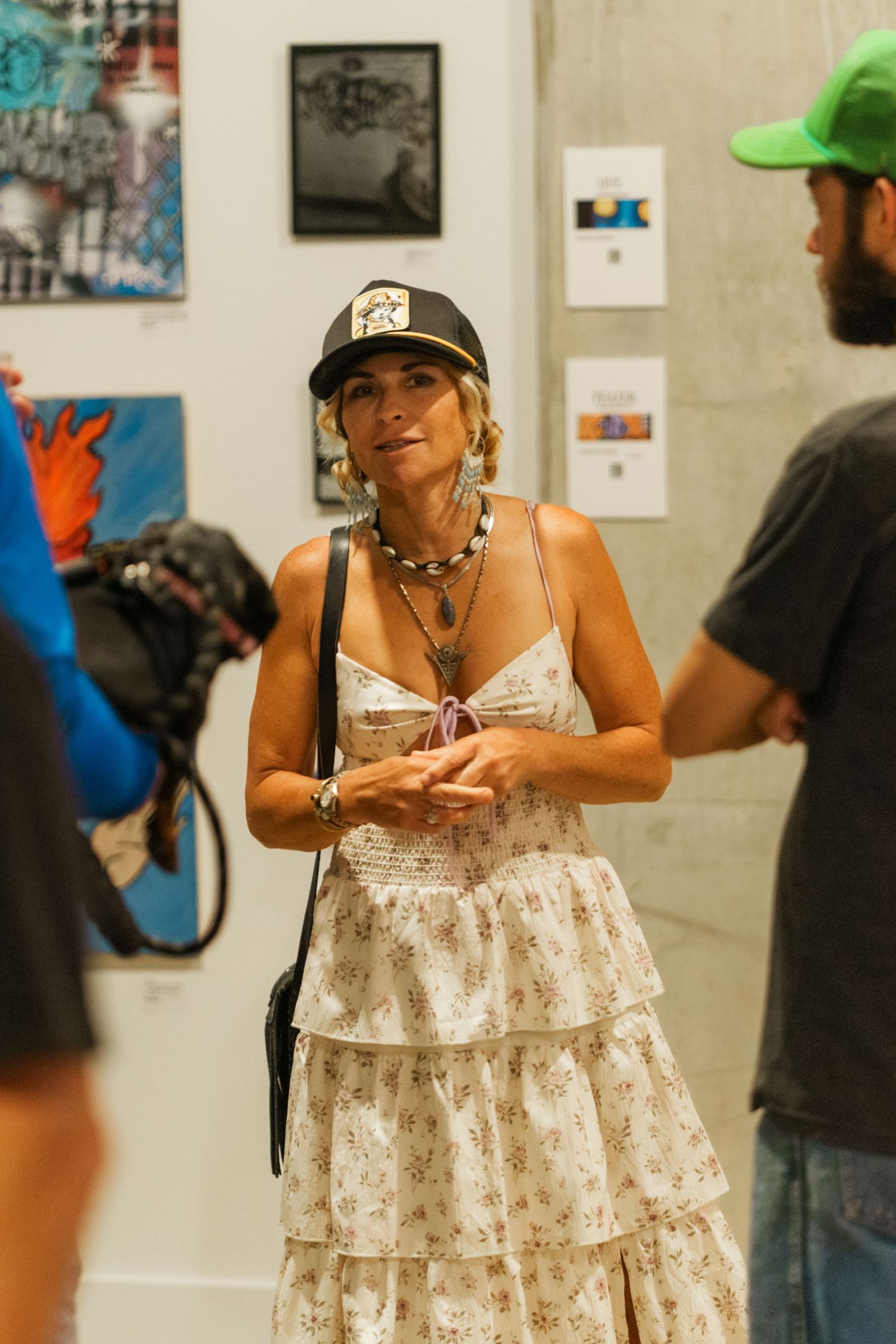 Woman in black cap and white floral dress engaging in conversation at an art gallery exhibition.