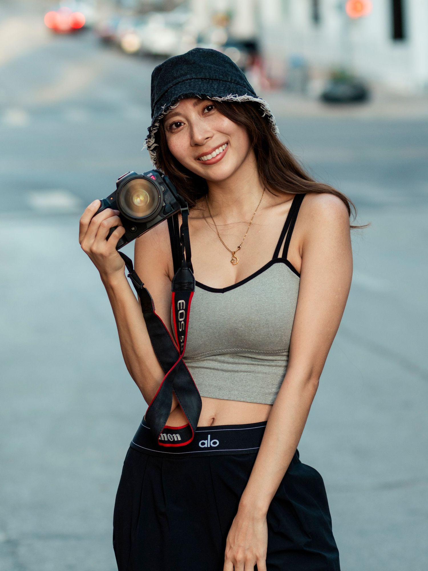 Smiling woman in crop top holding Canon camera outdoors on urban street.