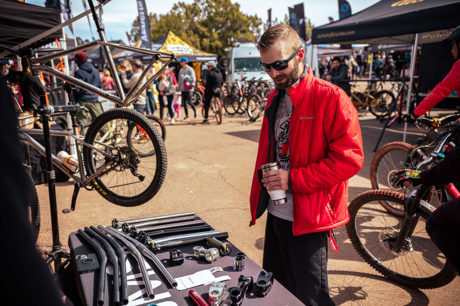 Man in red jacket examining bike components displayed on a table at an outdoor cycling event.