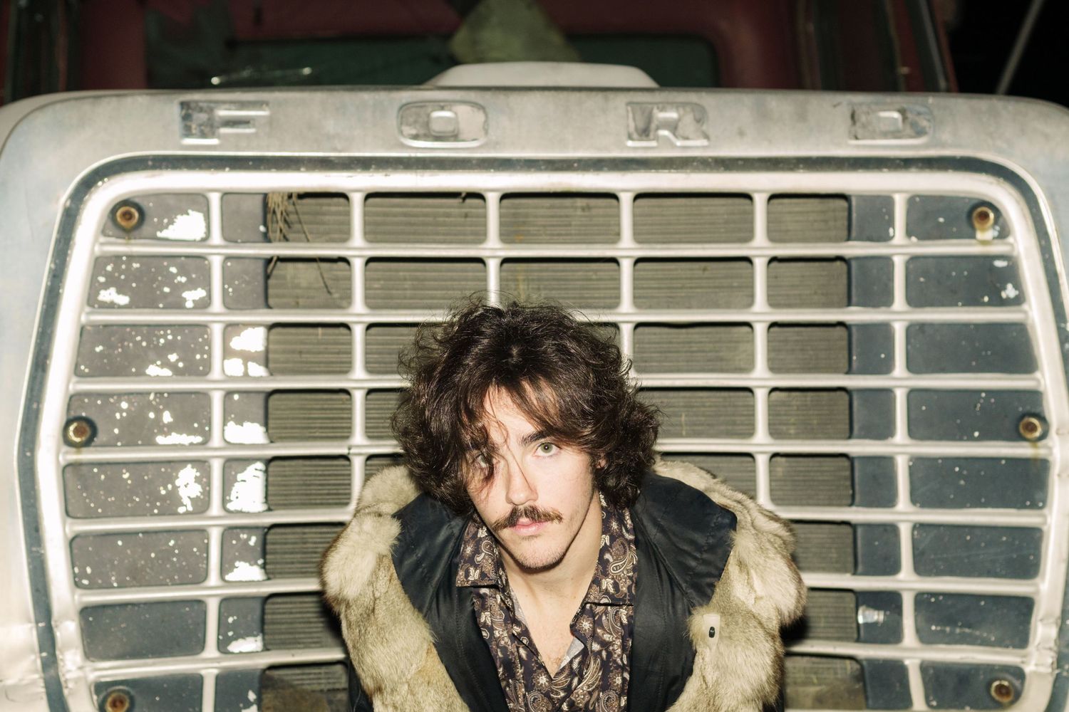 Curly-haired man in fur coat posing in front of vintage Ford truck grille.