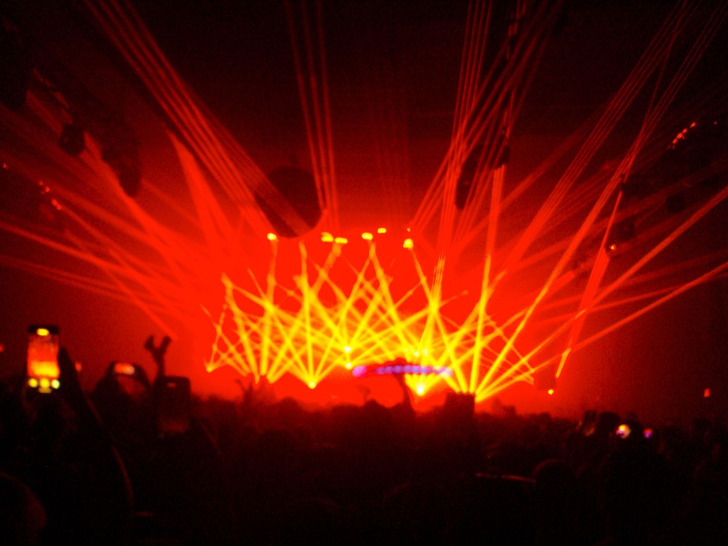 Red laser light show at a live concert with silhouetted crowd and dramatic beams radiating from the stage.