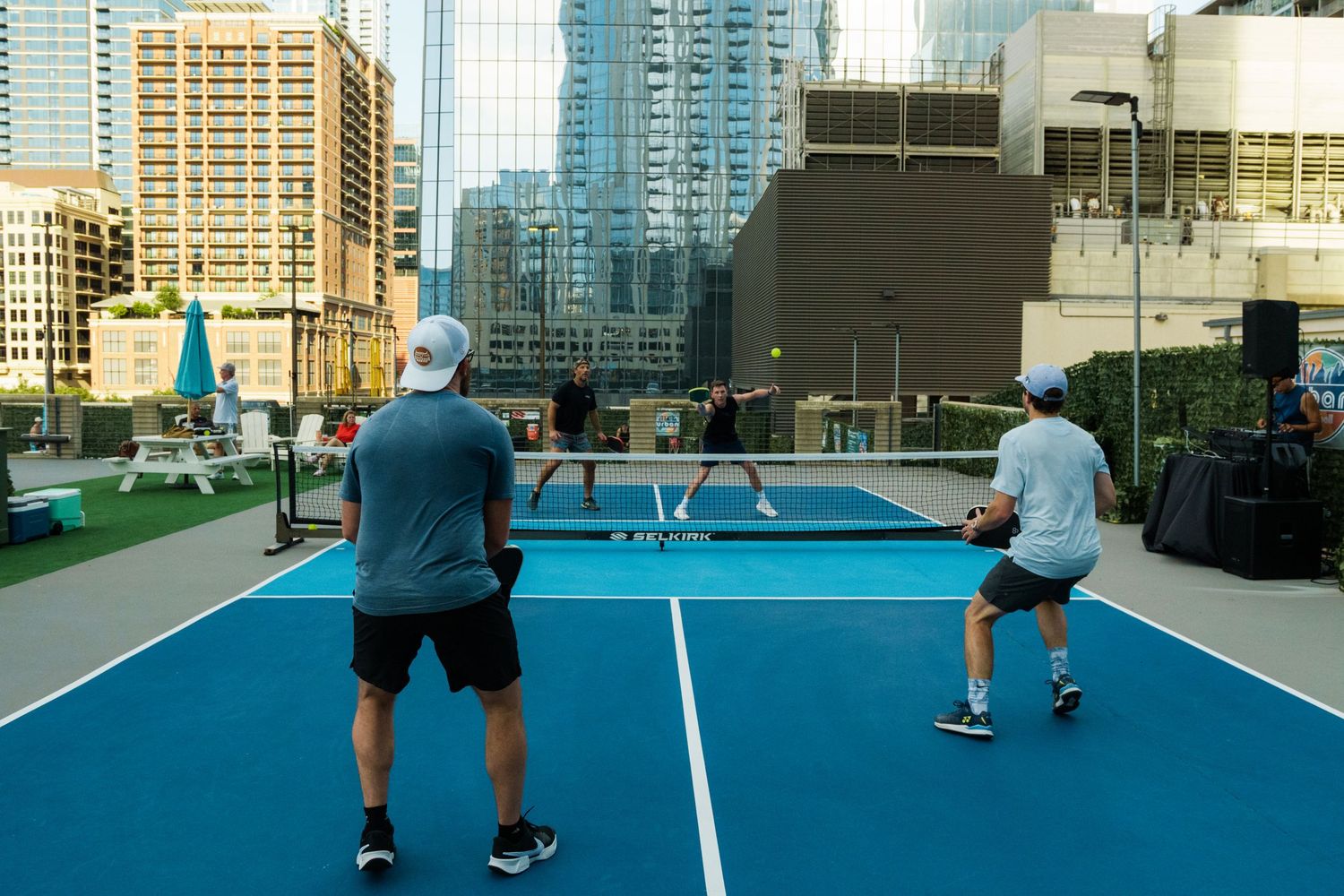 Four players enjoy a rooftop pickleball match on a blue court with city skyscrapers in the background.
