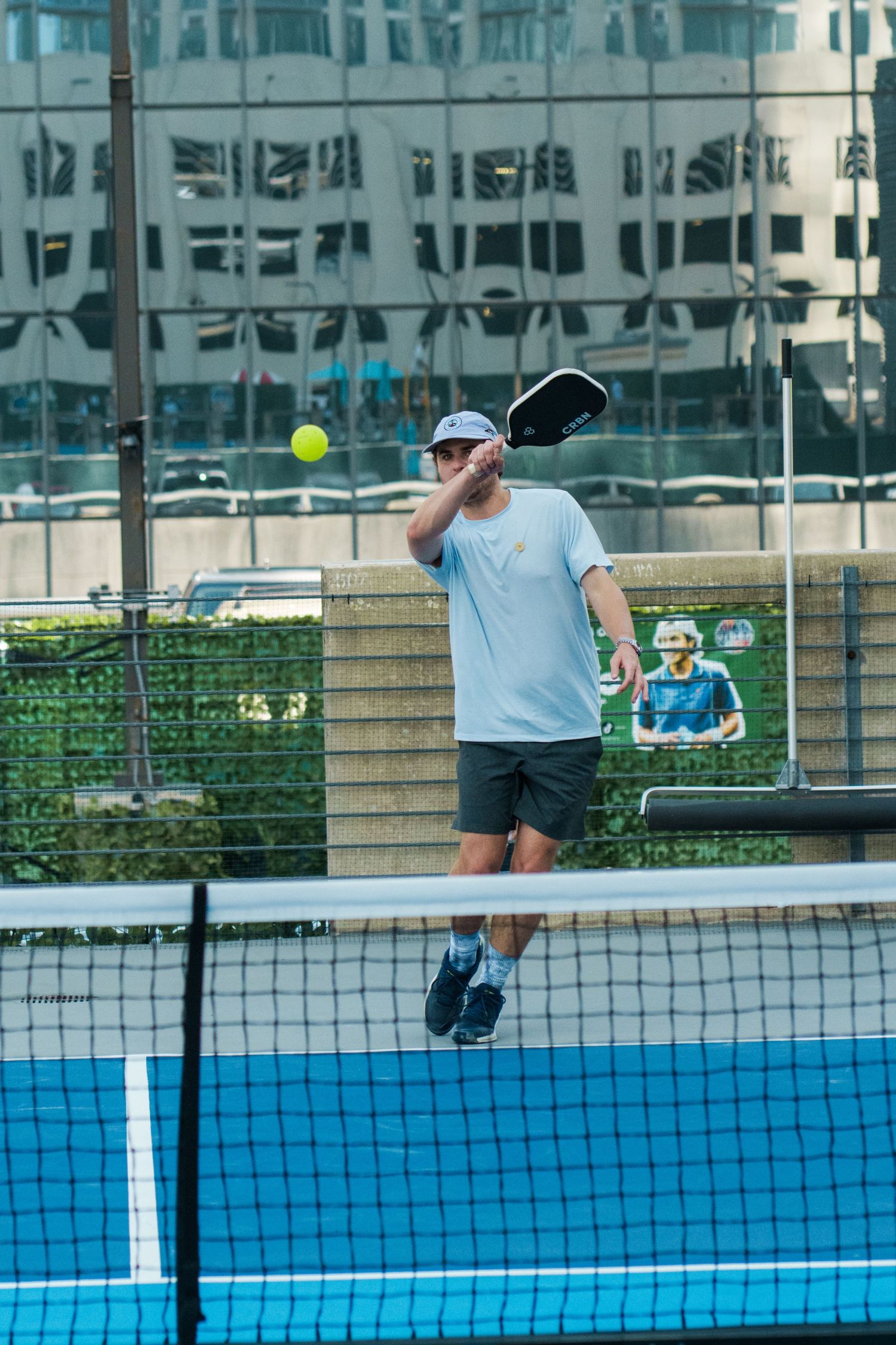 Man playing pickleball on an outdoor blue court, swinging paddle at yellow ball with urban buildings in background.